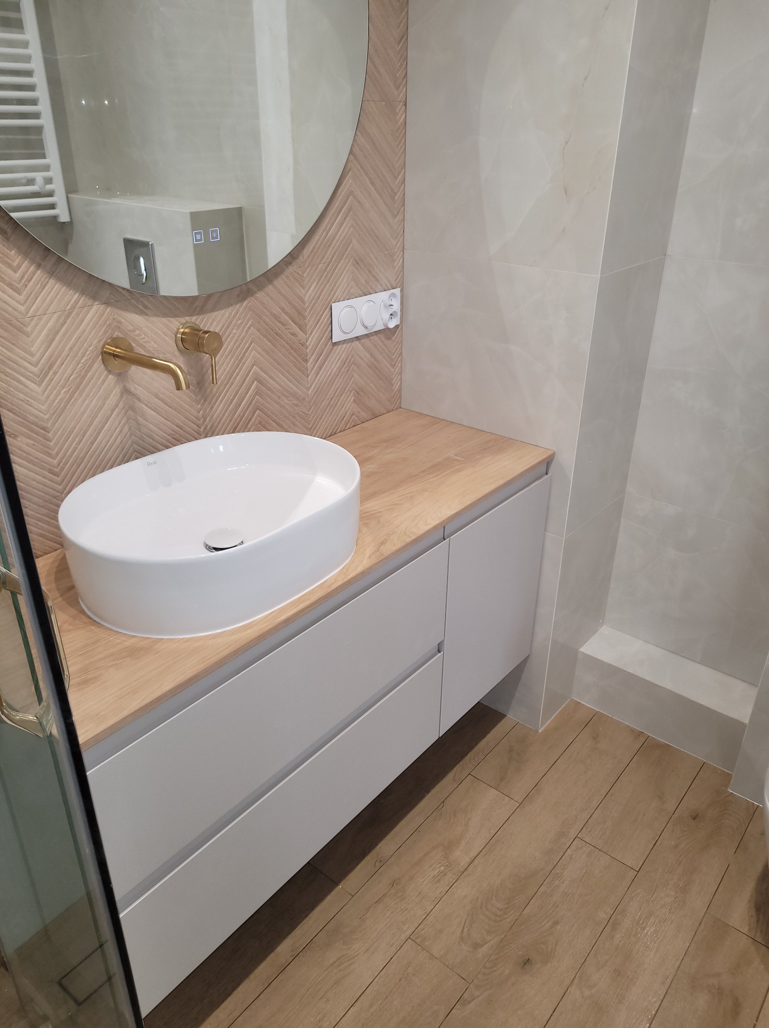 Nowoczesna łazienka z okrągłym lustrem, białą umywalką nablatową, złotą armaturą i drewnianą podłogą. Ściana z płytek ułożonych w jodełkę i beżowych płytek.