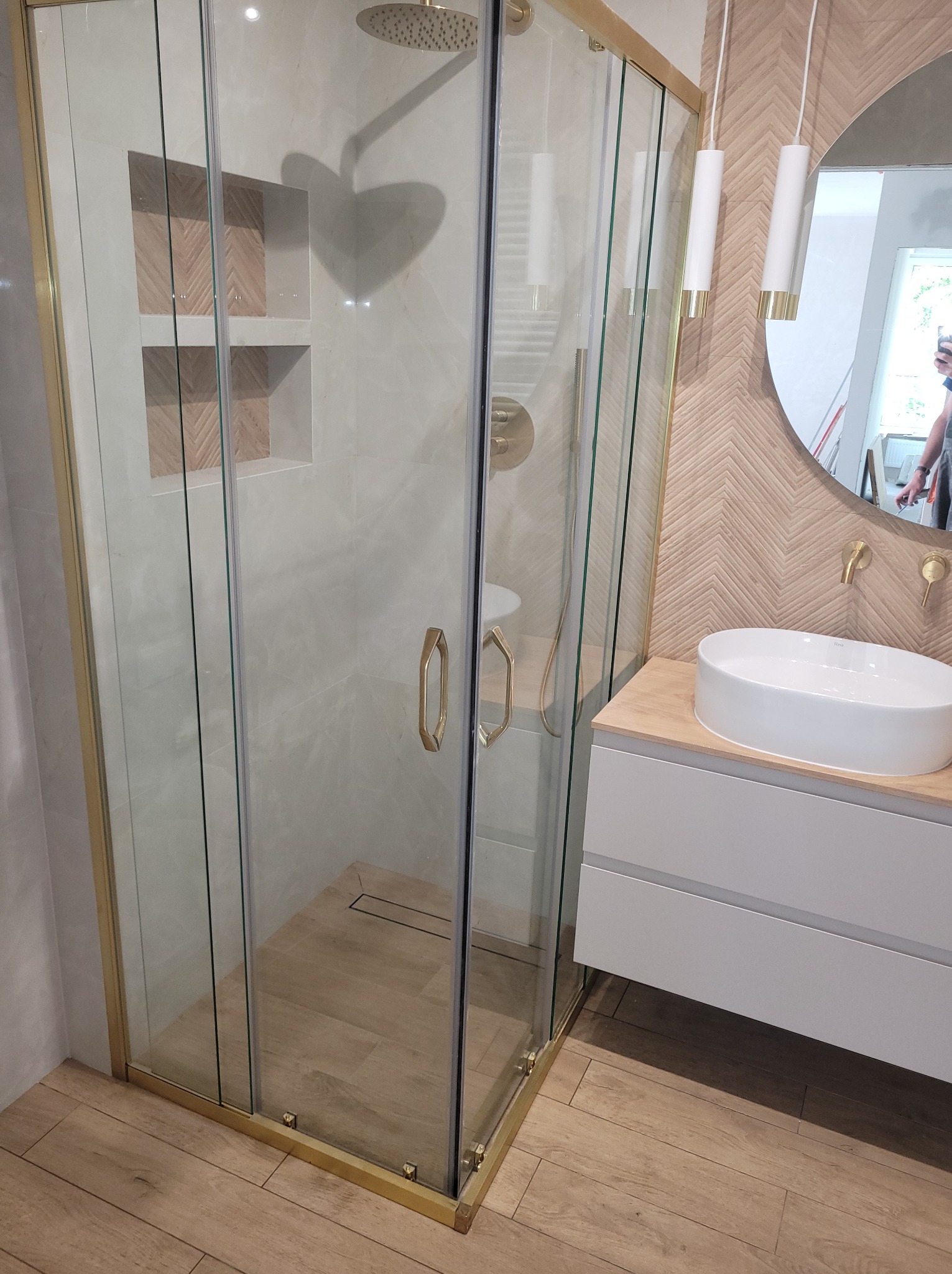 Nowoczesna łazienka: kabina prysznicowa ze złotymi profilami, umywalka nablatowa i lustro w drewnianej ramie. Widoczne oświetlenie punktowe i płytki imitujące drewno.