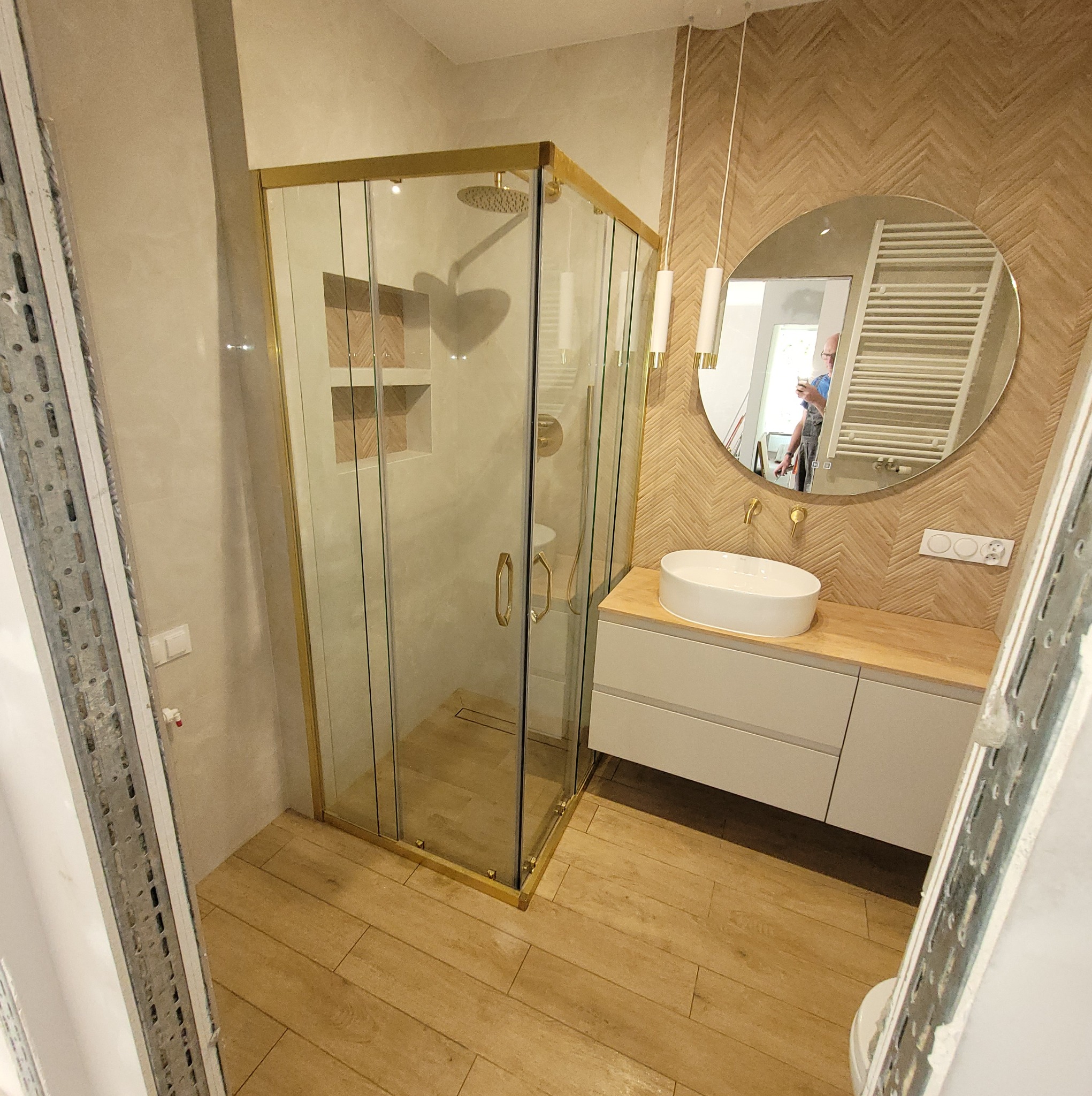 Nowoczesna łazienka z kabiną prysznicową w złotej ramie, okrągłym lustrem i umywalką nablatową na szafce z drewnianym blatem, wykończona płytkami imitującymi drewno.