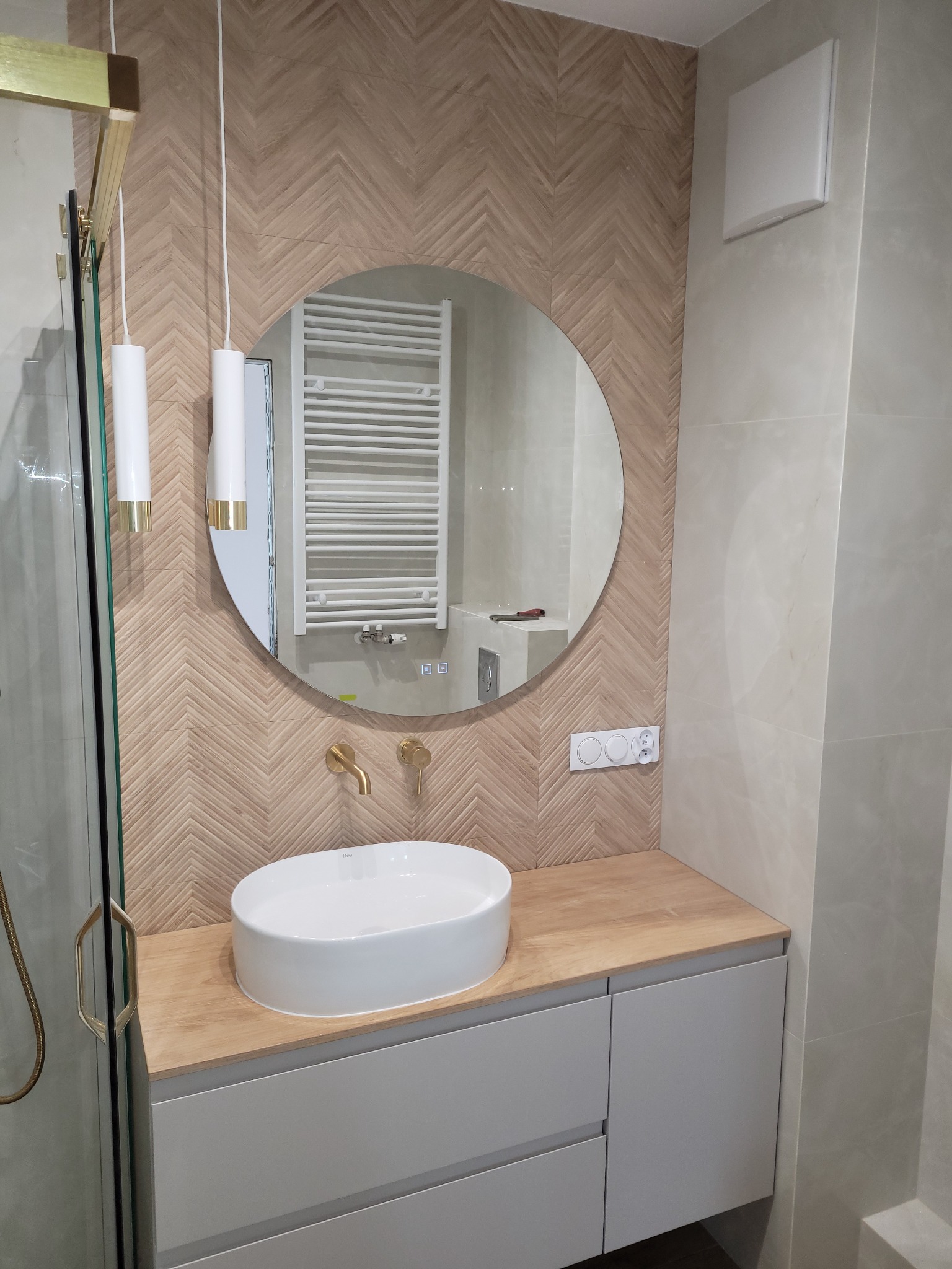 Nowoczesna łazienka z umywalką nablatową, okrągłym lustrem i drewnopodobną mozaiką na ścianie. Widoczna kabina prysznicowa z złotymi detalami i grzejnik drabinkowy w odbiciu lustra.