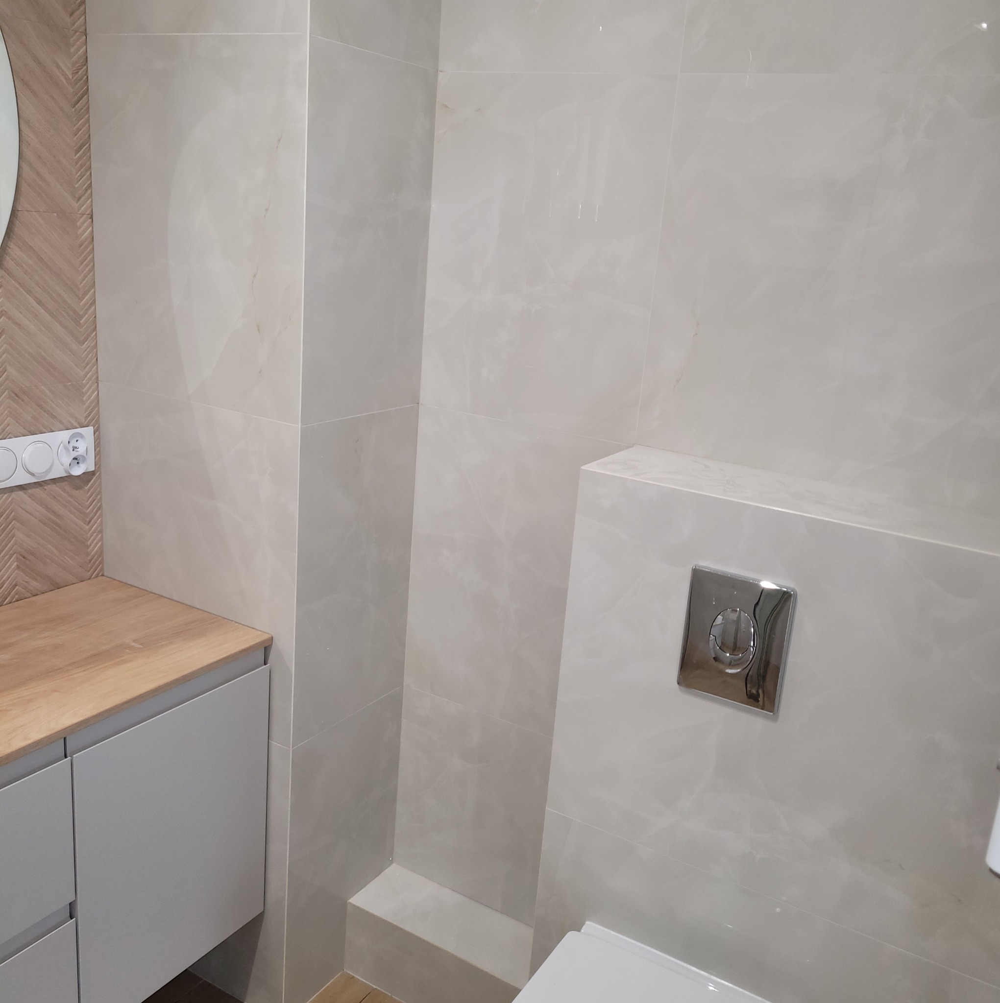 Jasna łazienka po remoncie: beżowe płytki imitujące marmur na ścianach, zabudowa WC z chromowanym przyciskiem, umywalka z szafką i drewnianym blatem.