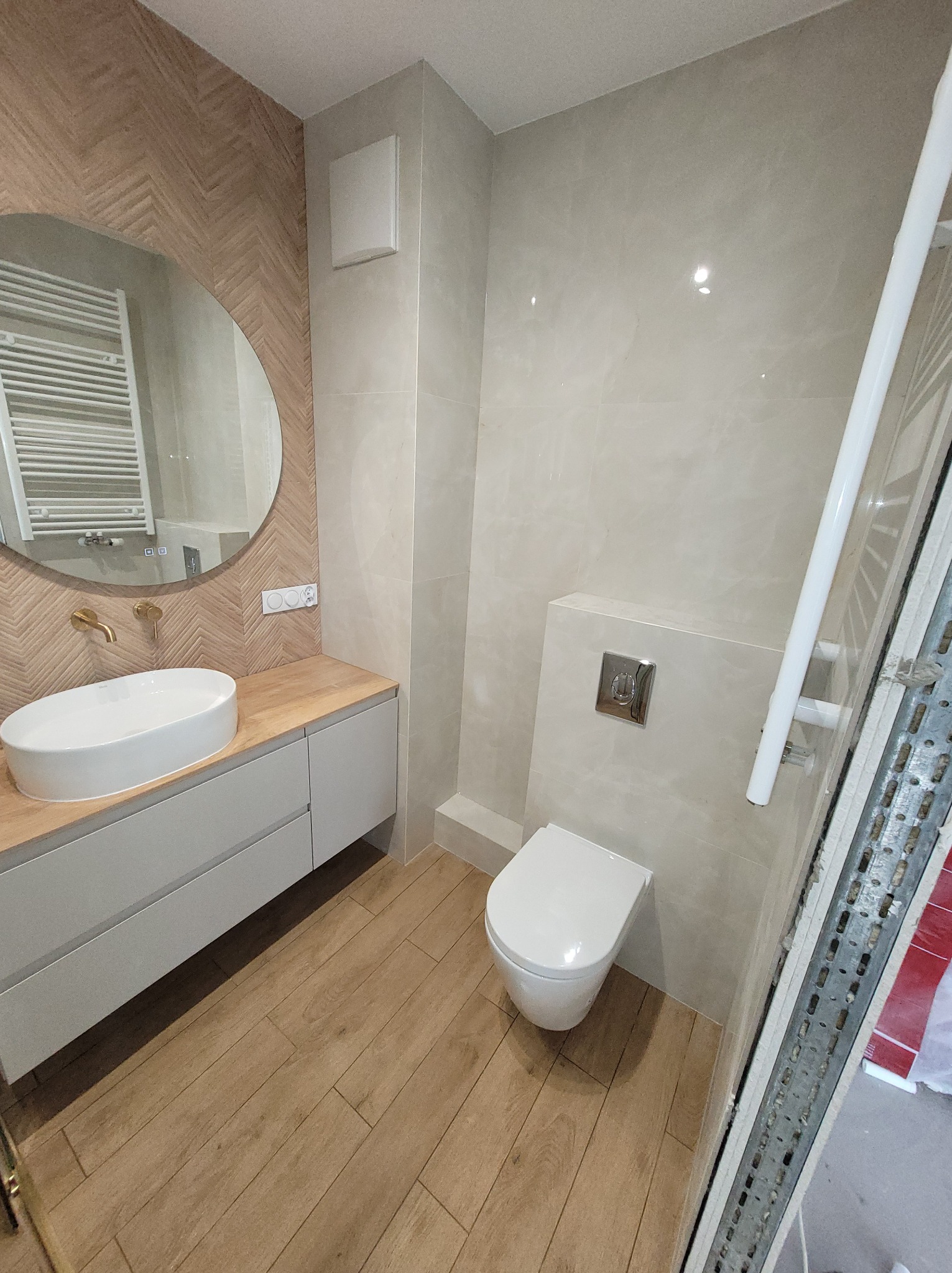 Jasna łazienka po remoncie: drewnopodobne płytki na podłodze i ścianie za umywalką, owalne lustro, podwieszana toaleta. Nowoczesny design.