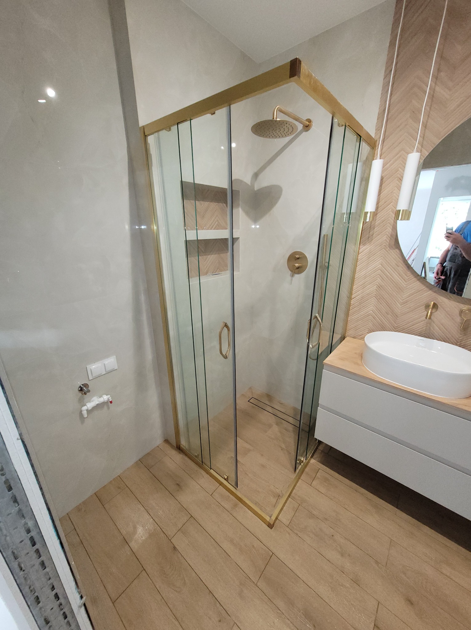 Elegancka łazienka z kabiną prysznicową w złotej ramie, drewnopodobne płytki na podłodze i ścianie, umywalka nablatowa i okrągłe lustro z oświetleniem.