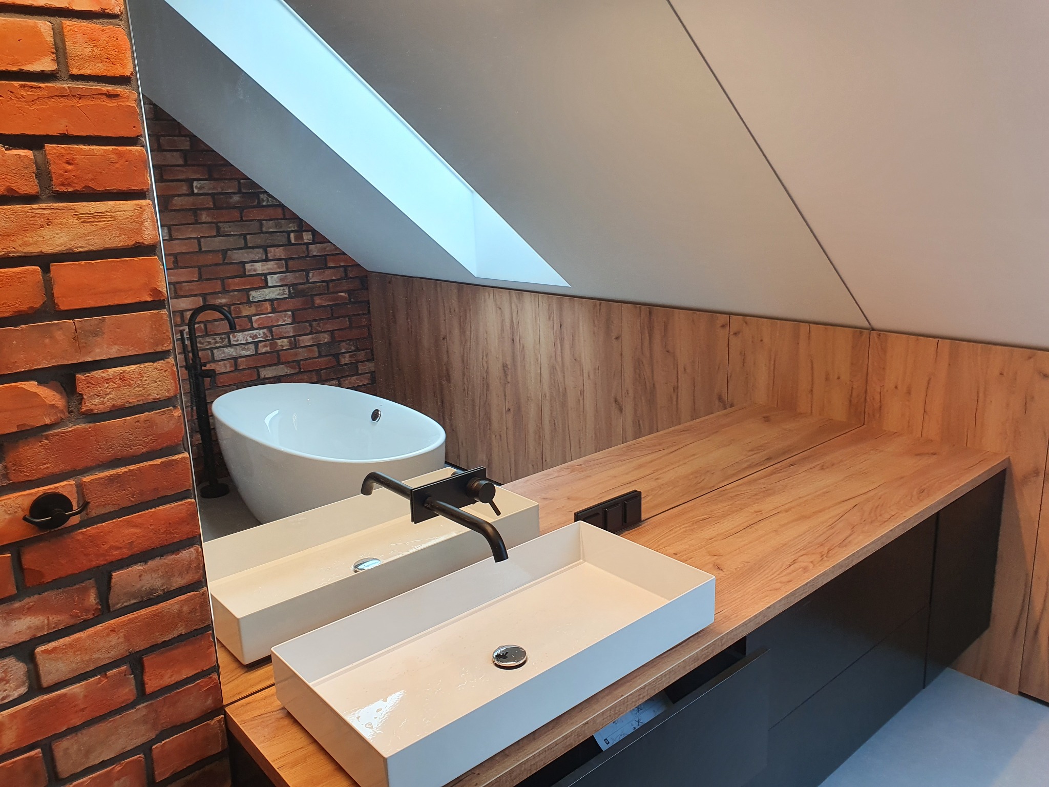 Nowoczesna łazienka na poddaszu: biała wanna wolnostojąca, dwie prostokątne umywalki nablatowe z czarną armaturą, ściana z cegły i drewniane panele ścienne.
