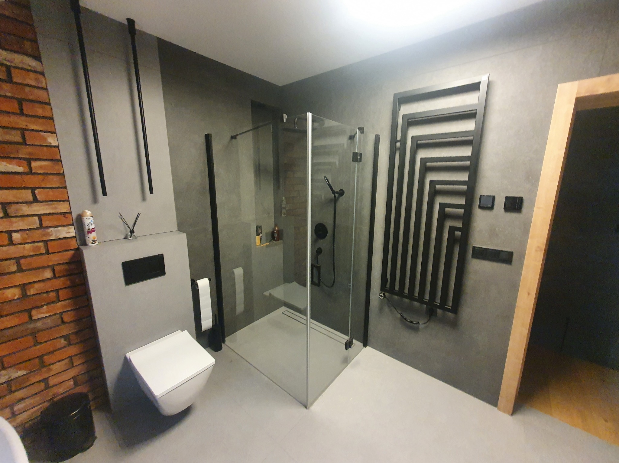 Nowoczesna łazienka w szarościach z kabiną prysznicową typu walk-in, wiszącą toaletą i grzejnikiem dekoracyjnym. Widoczna ściana z cegły i minimalistyczne dodatki.