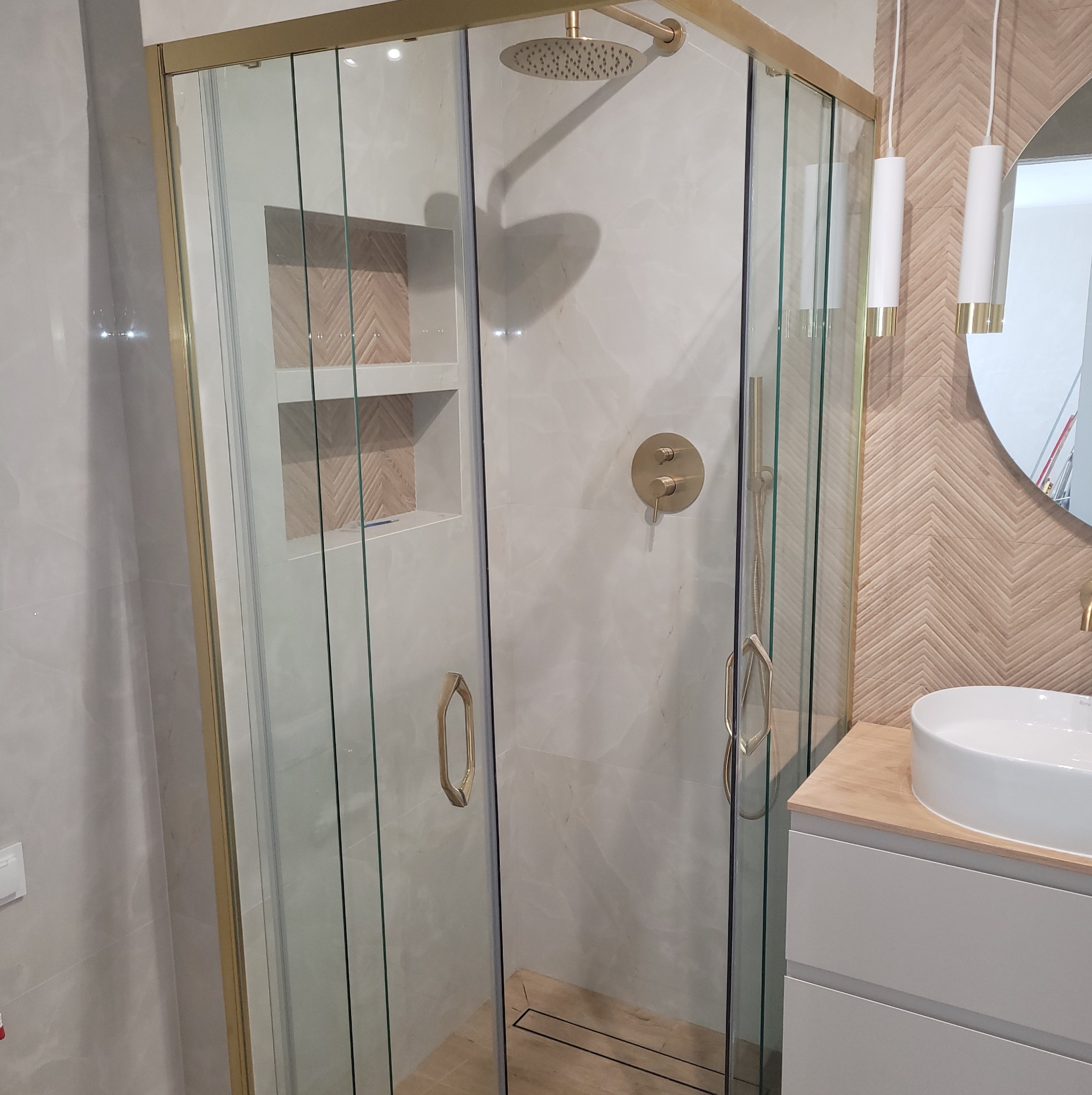 Nowoczesna kabina prysznicowa z złotymi detalami, wnęka z drewnianą mozaiką, umywalka nablatowa i lampy wiszące w wykończonej łazience. Jasne płytki, minimalistyczny design.