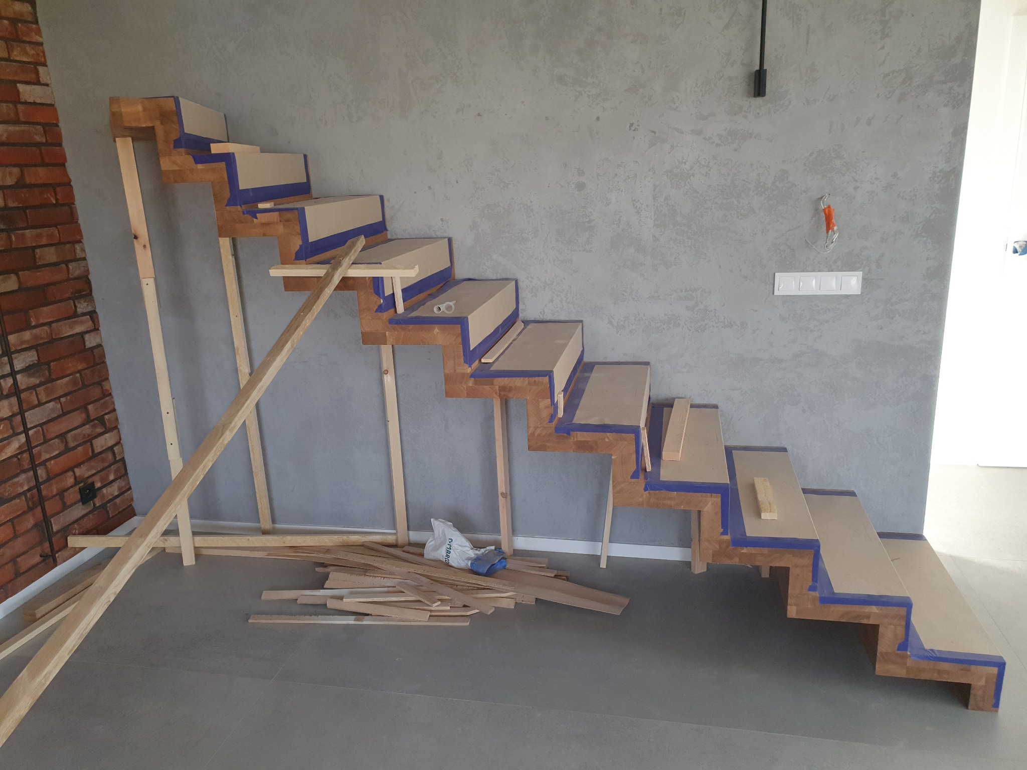 Montaż drewnianych schodów z zabezpieczeniem taśmą malarską, widoczne podpory i fragment ceglanej ściany. Szare tło ściany dodaje surowego charakteru.