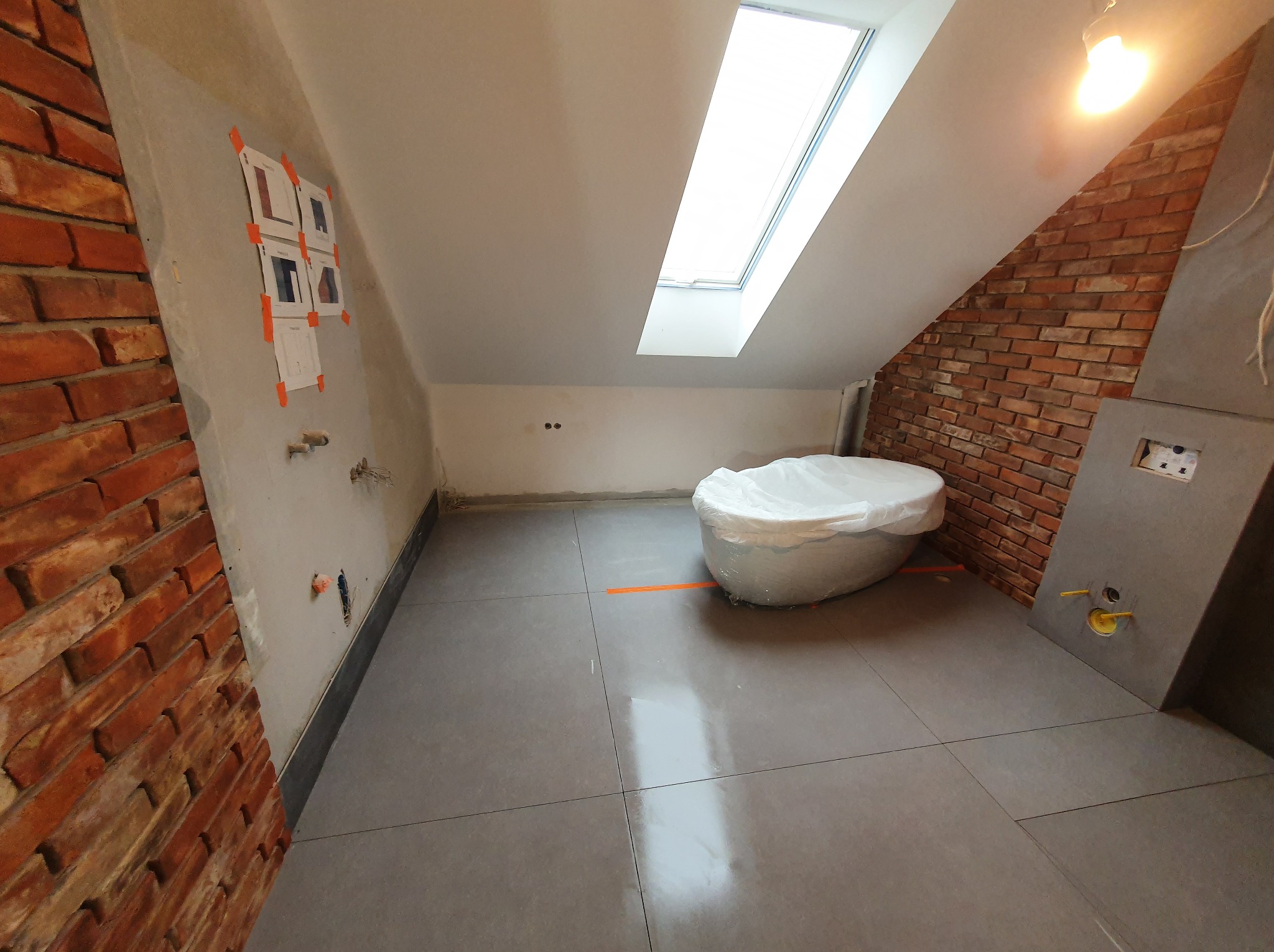 Łazienka na poddaszu w trakcie prac: szare płytki, cegła na ścianie, okno dachowe i wanna zabezpieczona folią. Widoczne przygotowania do montażu armatury.