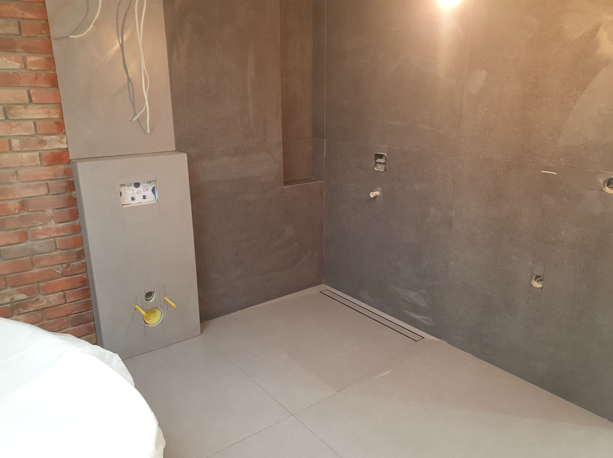 Szara łazienka w trakcie wykańczania: płytki na ścianach, zabudowa WC, odpływ liniowy w podłodze, fragment ściany z cegły. Widoczne przygotowania do instalacji.