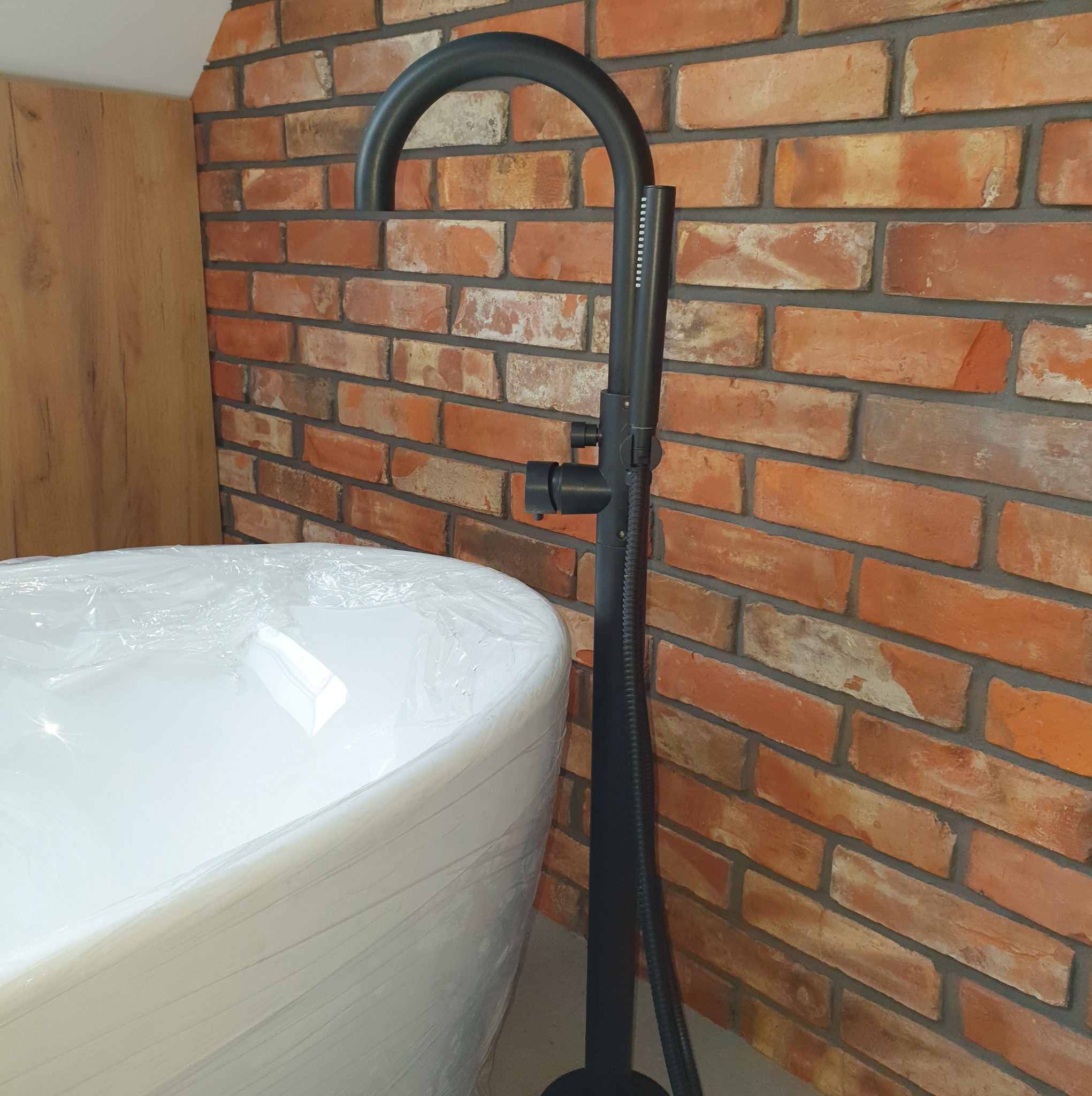 Nowoczesna, czarna bateria wannowa wolnostojąca na tle ściany z cegły. Wanna owinięta folią ochronną. Element wykończenia łazienki w stylu industrialnym.