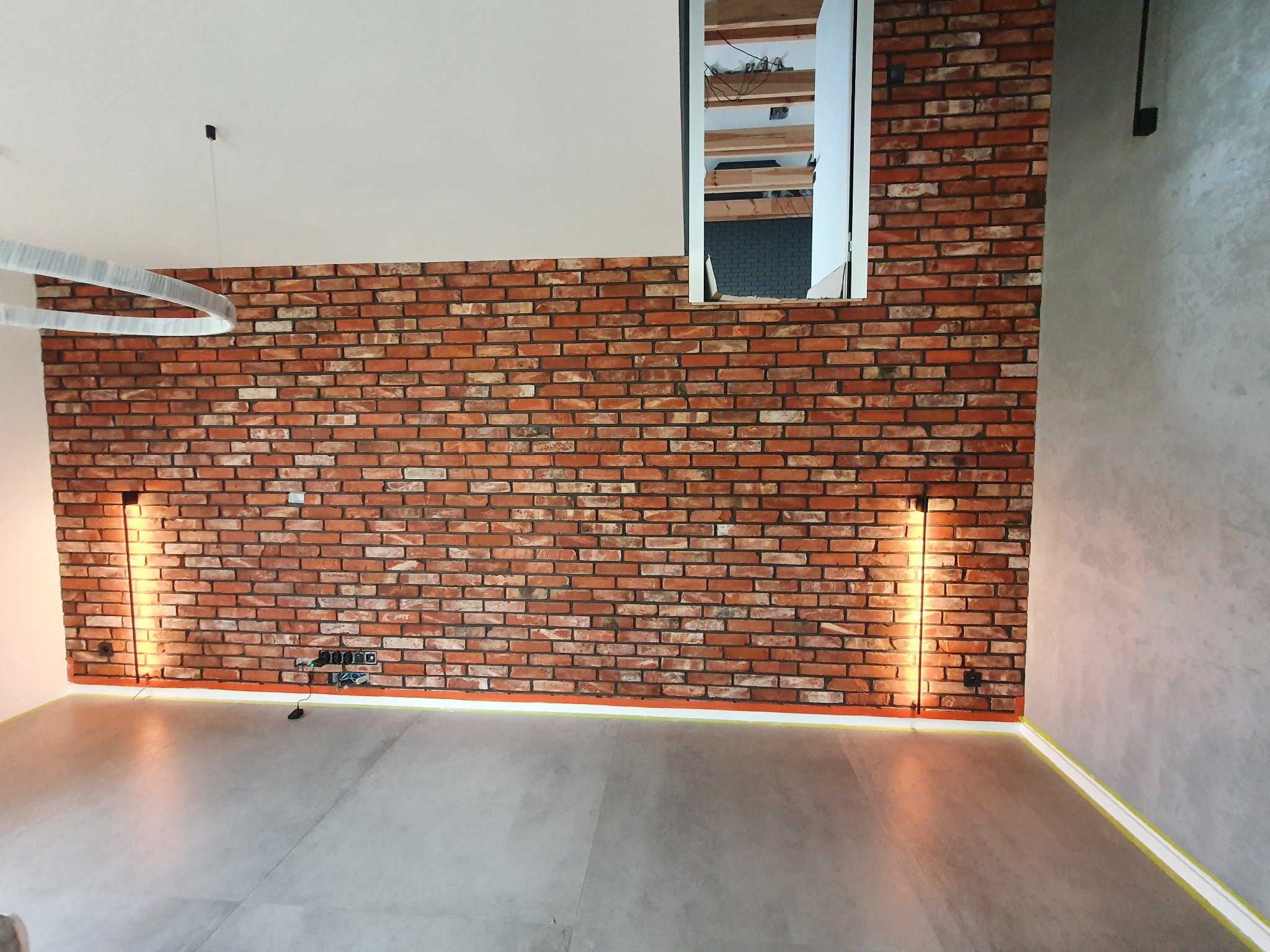 Ściana z cegły klinkierowej z oświetleniem punktowym, szara podłoga i fragment surowej ściany. Widoczne gniazdka elektryczne i taśmy malarskie na podłodze.