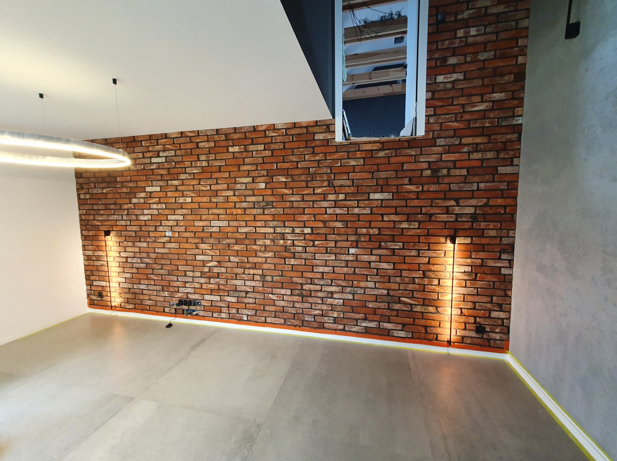 Ceglana ściana z oświetleniem LED w nowocześnie wykończonym wnętrzu. Widoczne szare płytki podłogowe i fragment okna. Taśma malarska zabezpiecza krawędzie.