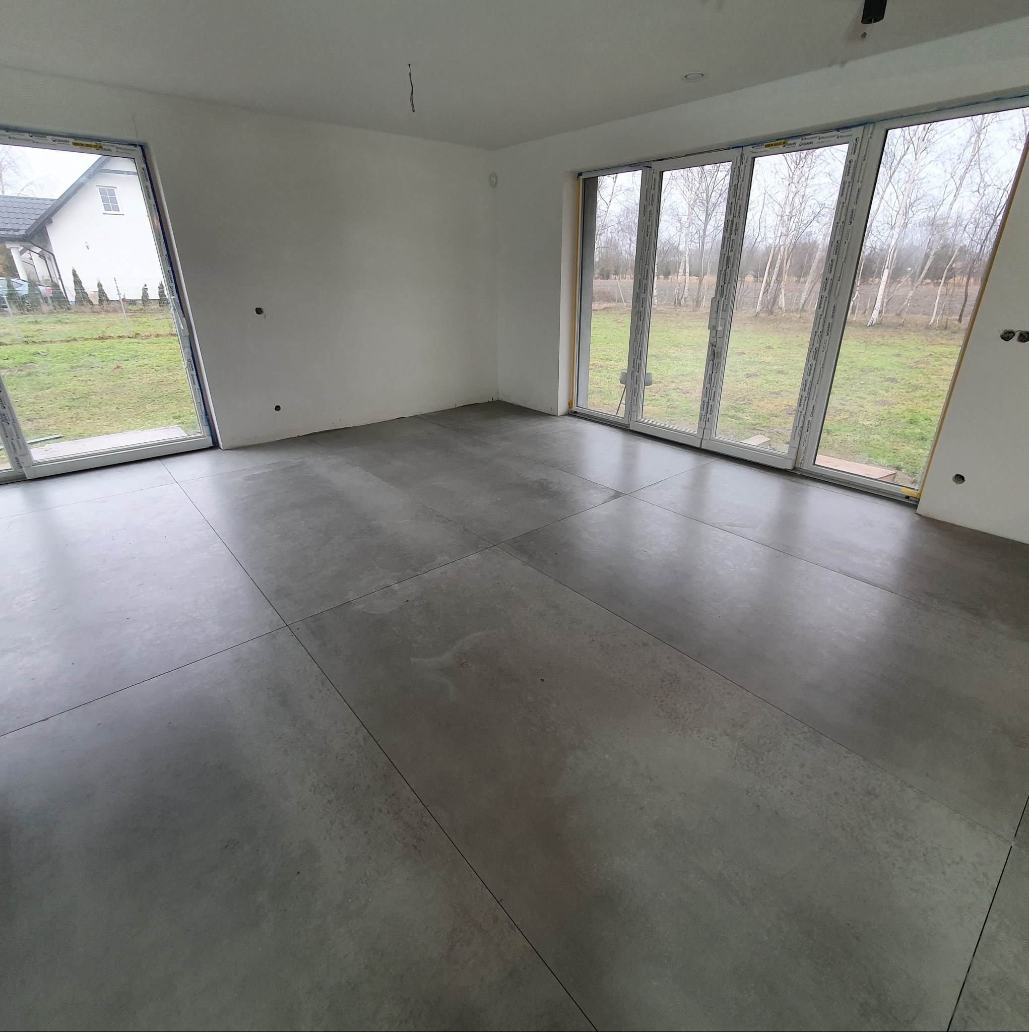 Szare płytki podłogowe w jasnym, przestronnym pokoju z dużymi oknami w Wejherowie. Widok na zewnątrz, świeżo pomalowane ściany, surowy stan wykończenia.