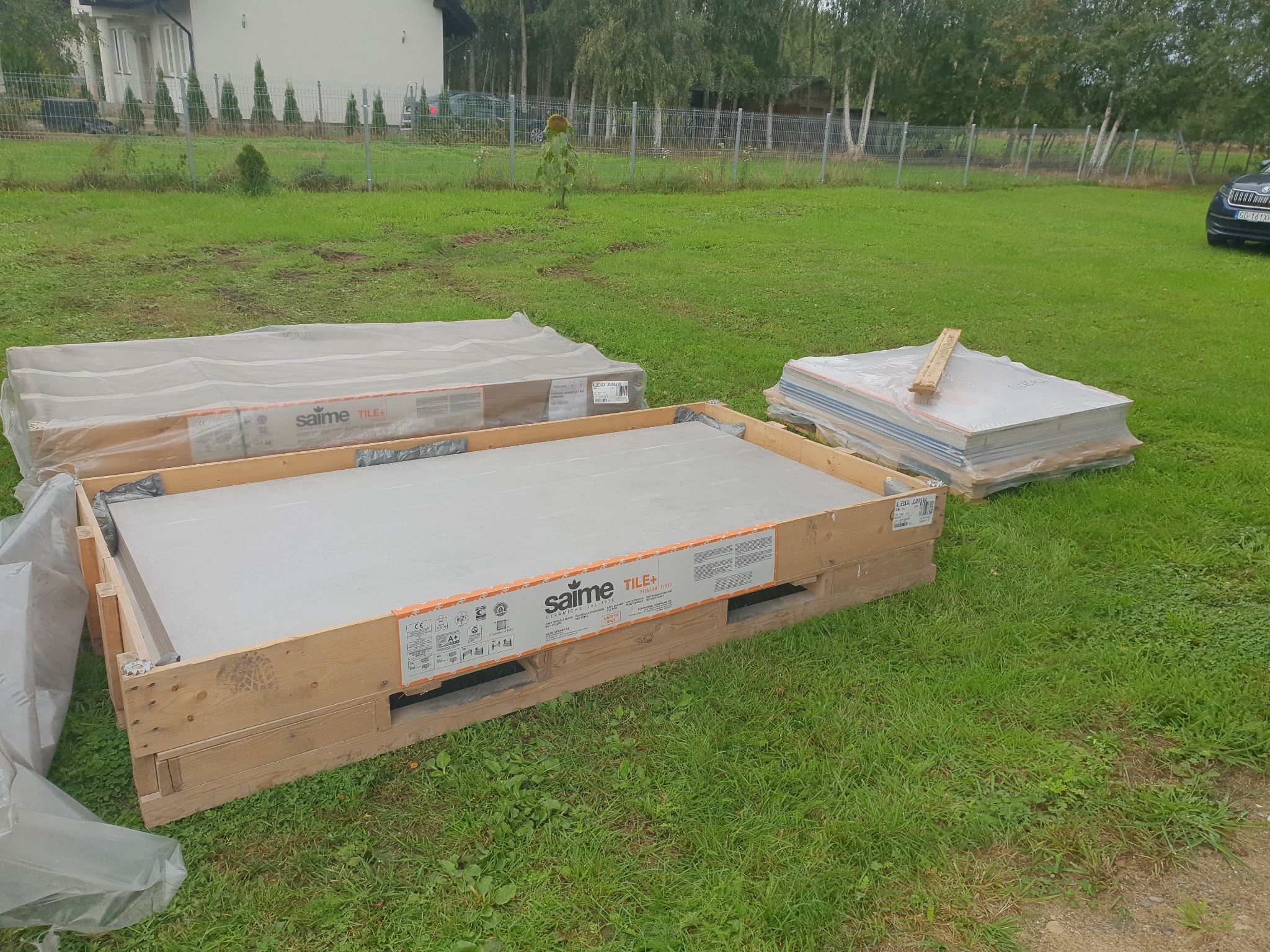 Paczki z płytami marki Saime Tile+ leżą na trawie przed domem. Widoczne palety i opakowania z szarymi płytami. W tle zieleń trawnika i budynek mieszkalny.