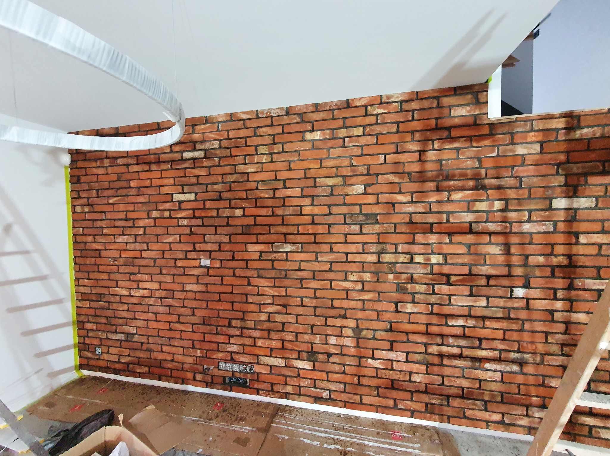 Ściana z cegły klinkierowej w trakcie montażu, widoczne puszki elektryczne, zabezpieczona podłoga i fragment drabiny. Oświetlenie ledowe na suficie.