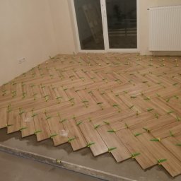 VIKTOR PRODANIUK VIKTORLUXPRO - Układanie drewnopodobnych płytek podłogowych w jodełkę, z użyciem zielonych krzyżyków dystansowych, widoczny poziomica i resztki kleju.