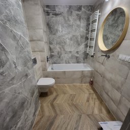 VIKTOR PRODANIUK VIKTORLUXPRO - Nowoczesna łazienka po remoncie: szare płytki imitujące marmur na ścianach, drewnopodobna podłoga ułożona w jodełkę, biała wanna i toaleta, okrągłe lustro z drewnianą ramą, grzejnik drabinkowy.