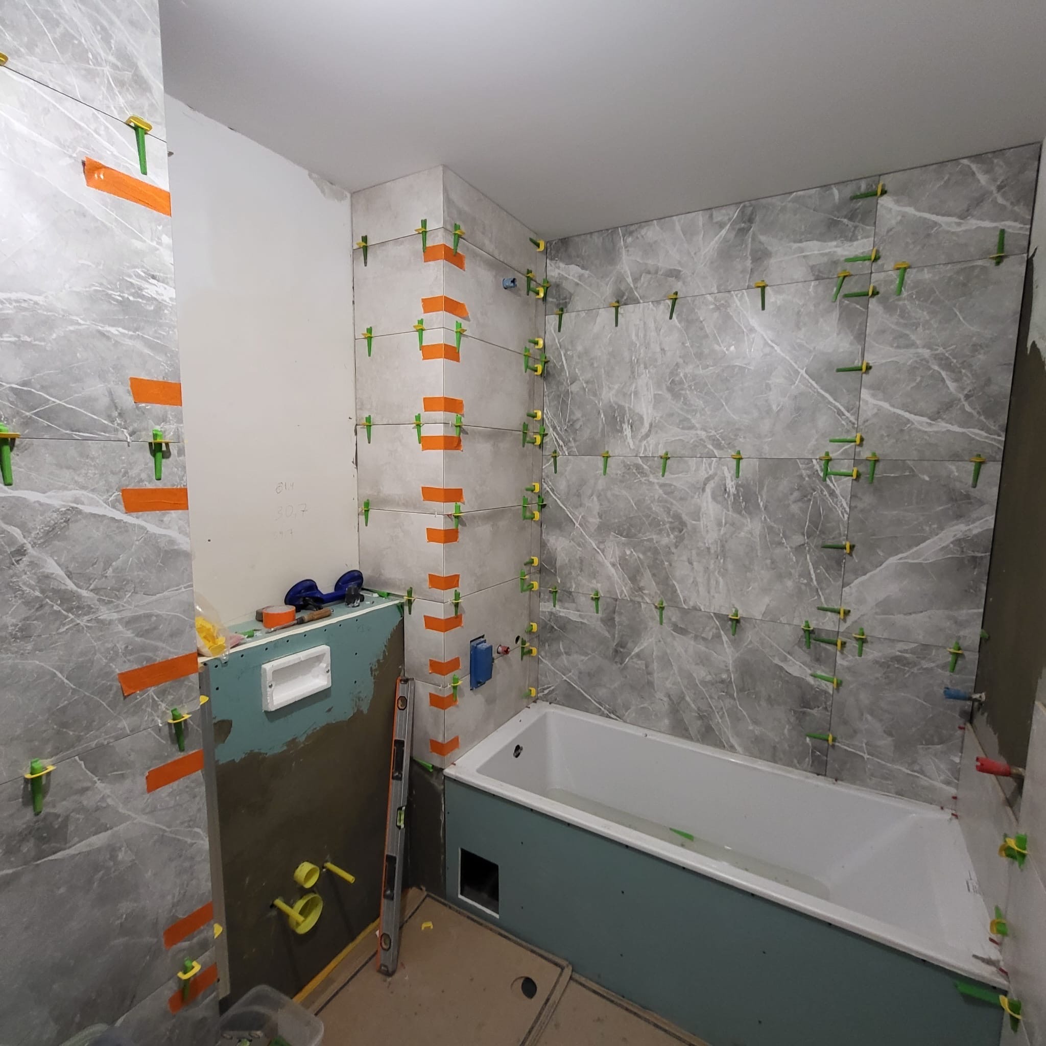 Układanie szarych płytek imitujących marmur w łazience we Wrocławiu, widoczne pomarańczowe paski i zielone kliny do poziomowania, przygotowana obudowa wanny z płyty gipsowej.