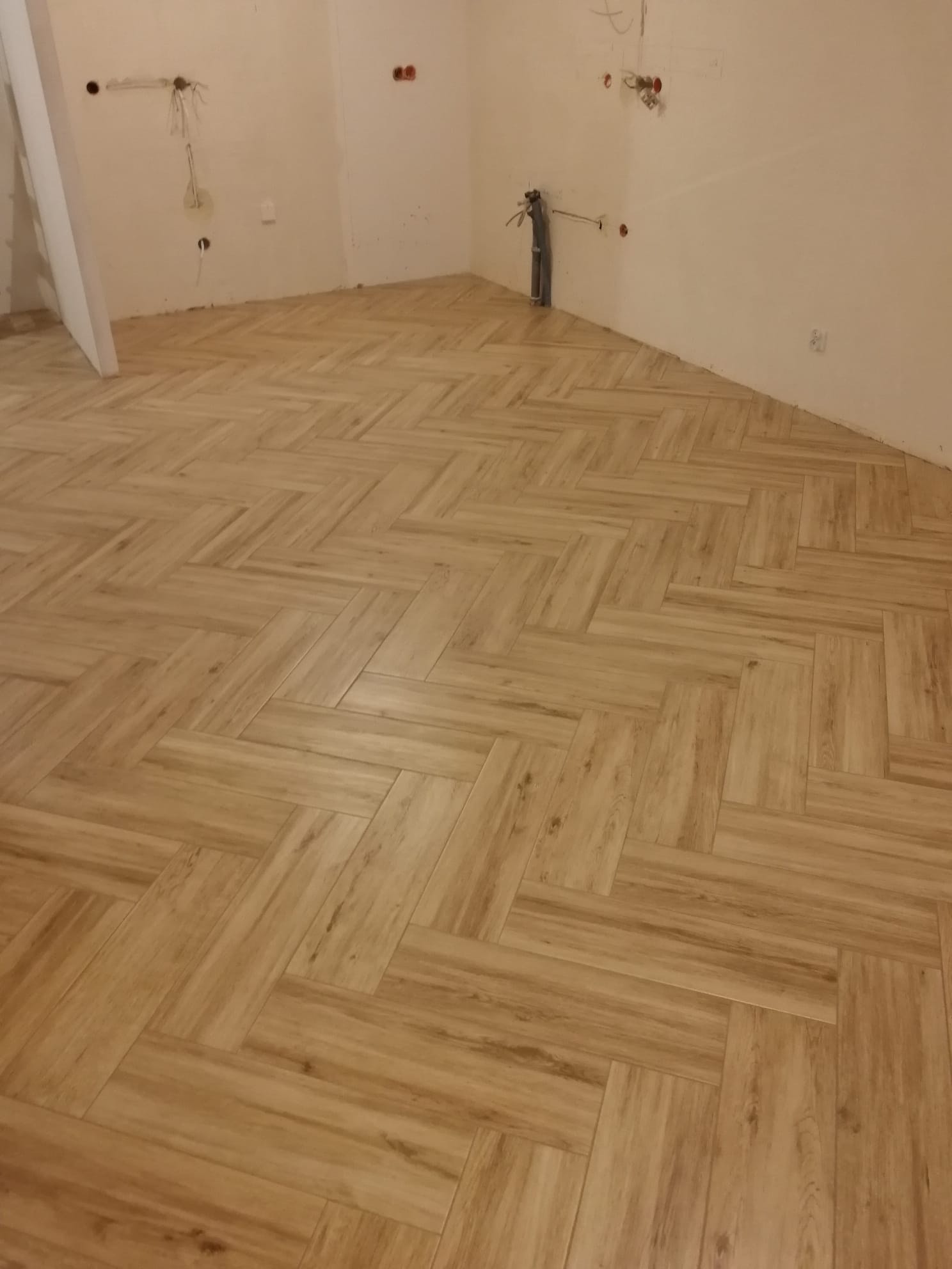 Ułożona podłoga z płytek imitujących drewno w jodełkę, widoczne wyprowadzenia instalacji na ścianach w surowym stanie.