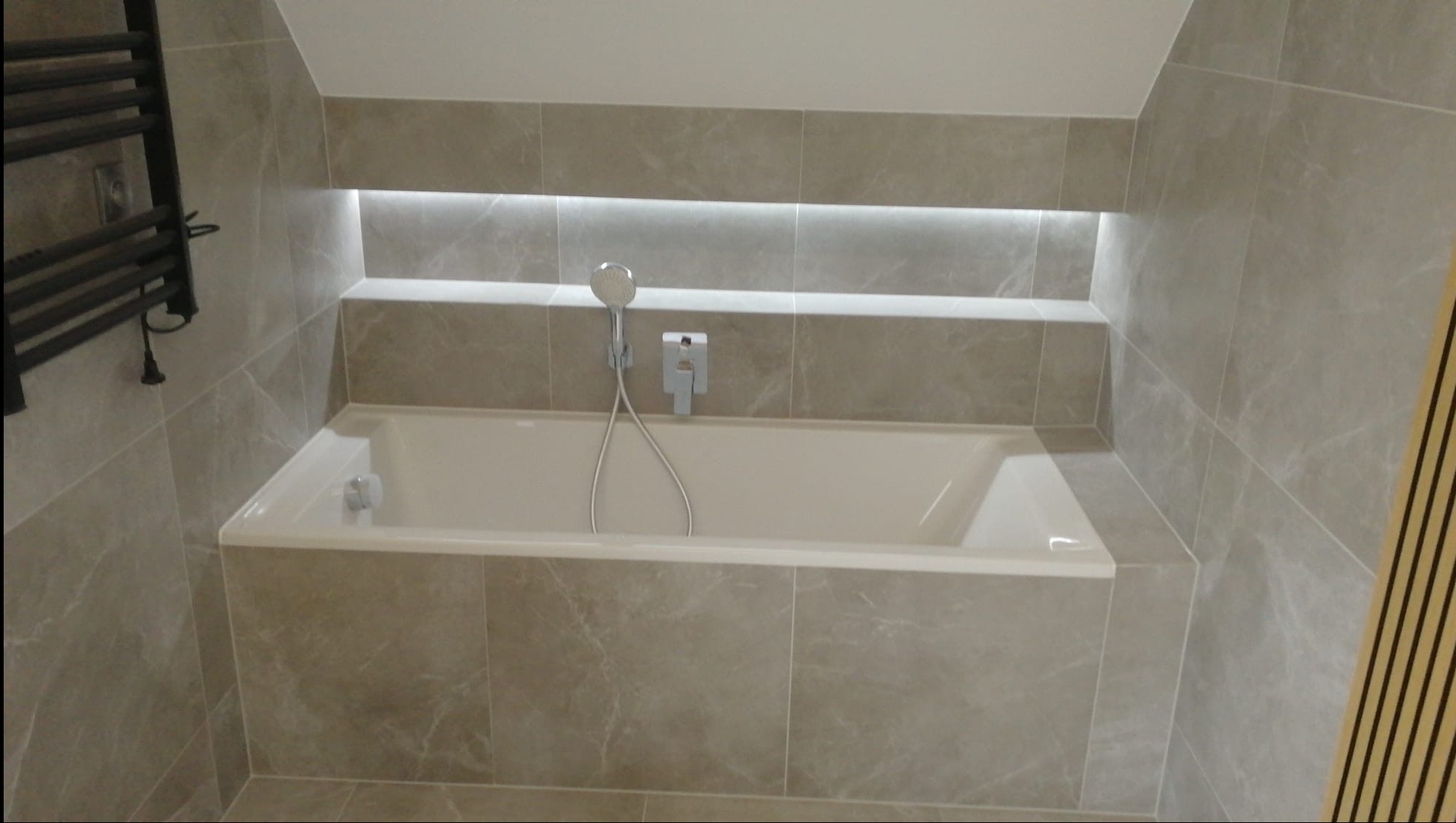 Nowoczesna łazienka z wanną obudowaną płytkami imitującymi marmur, podświetlana wnęka nad wanną, grzejnik drabinkowy po lewej stronie.