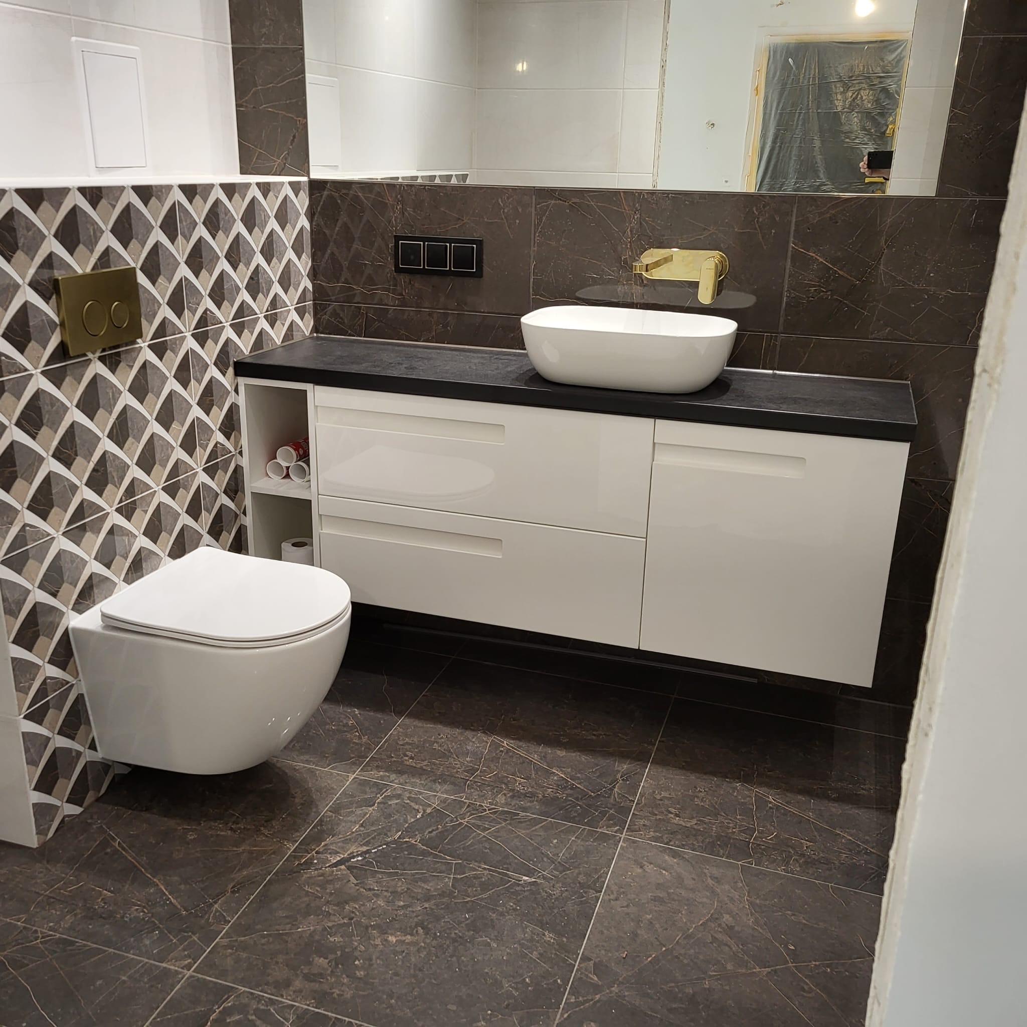 Nowoczesna łazienka z wiszącą toaletą, białą umywalką nablatową na czarnym blacie i brązowymi płytkami imitującymi marmur, z elementami złotej armatury i mozaiki.