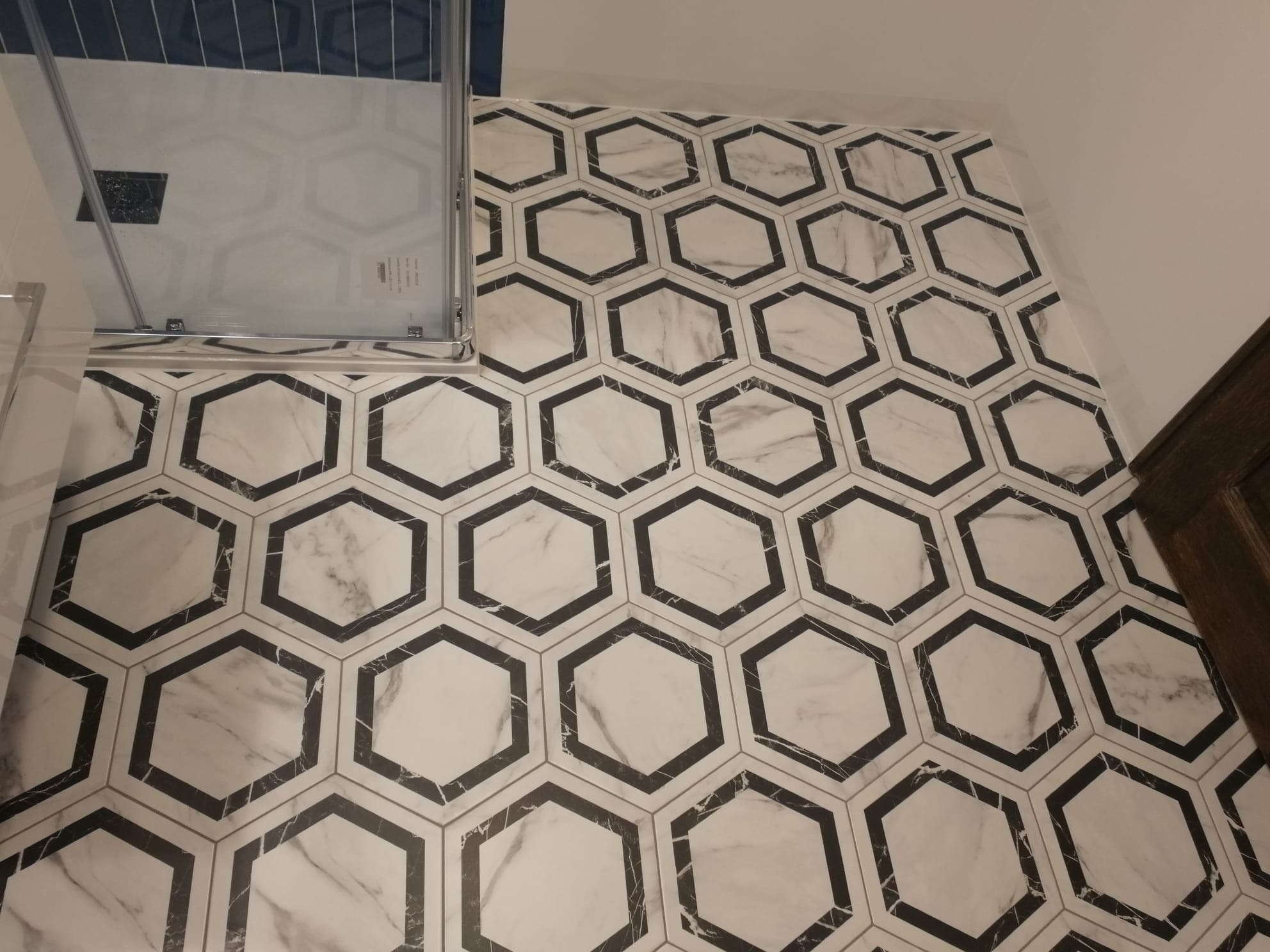 Ułożona podłoga z heksagonalnych płytek imitujących marmur, z czarnymi obwódkami, widok z góry na fragment kabiny prysznicowej z przezroczystego szkła.