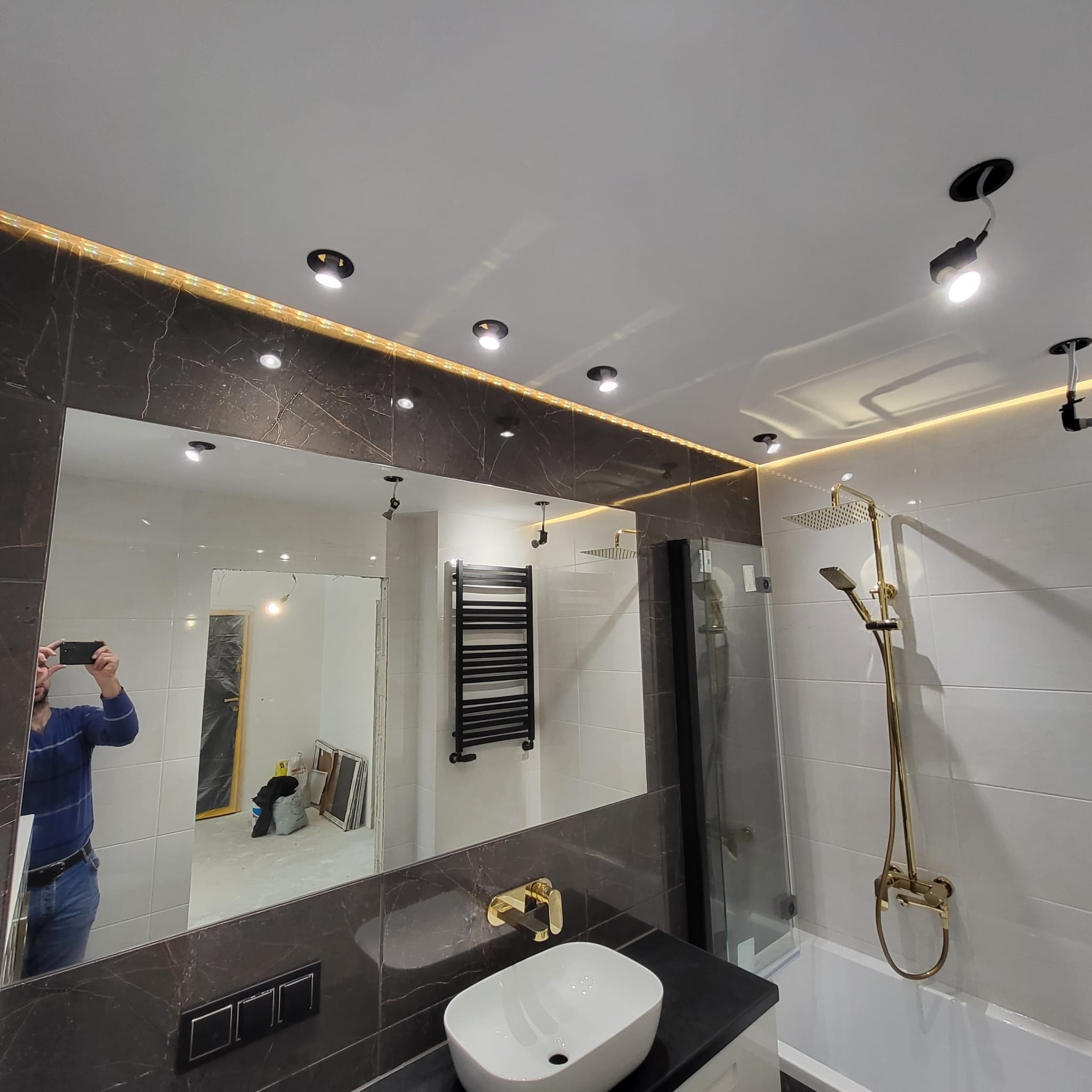 Nowoczesna łazienka z marmurowymi płytkami, złotą armaturą i podświetlanym lustrem, widok na kabinę prysznicową i grzejnik drabinkowy, odbicie osoby robiącej zdjęcie.