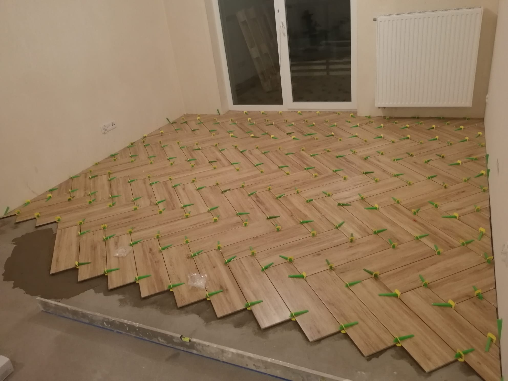 Układanie drewnopodobnych płytek podłogowych w jodełkę, z użyciem zielonych krzyżyków dystansowych, widoczny poziomica i resztki kleju.
