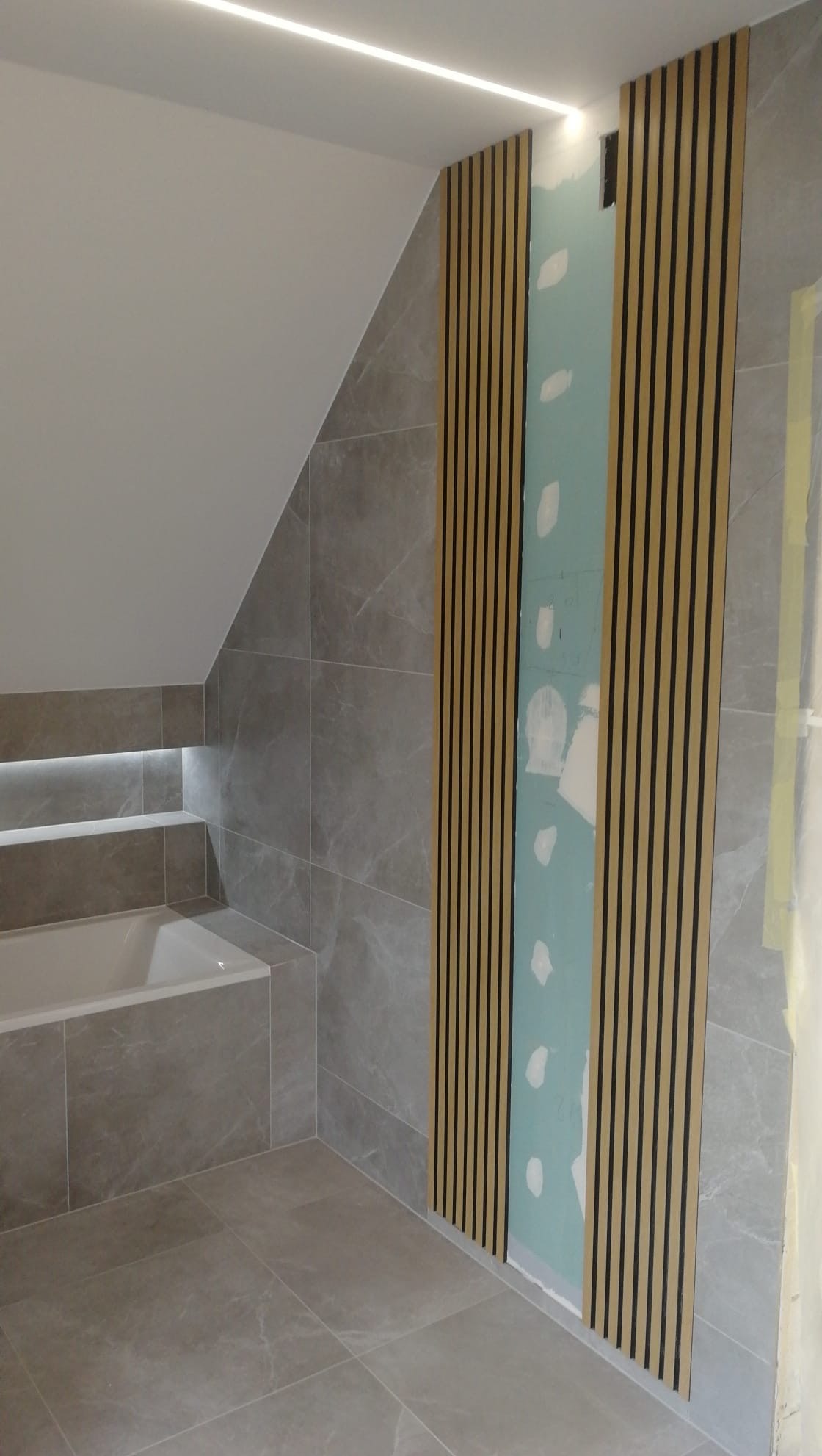 Szara łazienka z podświetlaną wanną i panelami ściennymi z listew, widoczna płyta gipsowo-kartonowa przed montażem paneli.