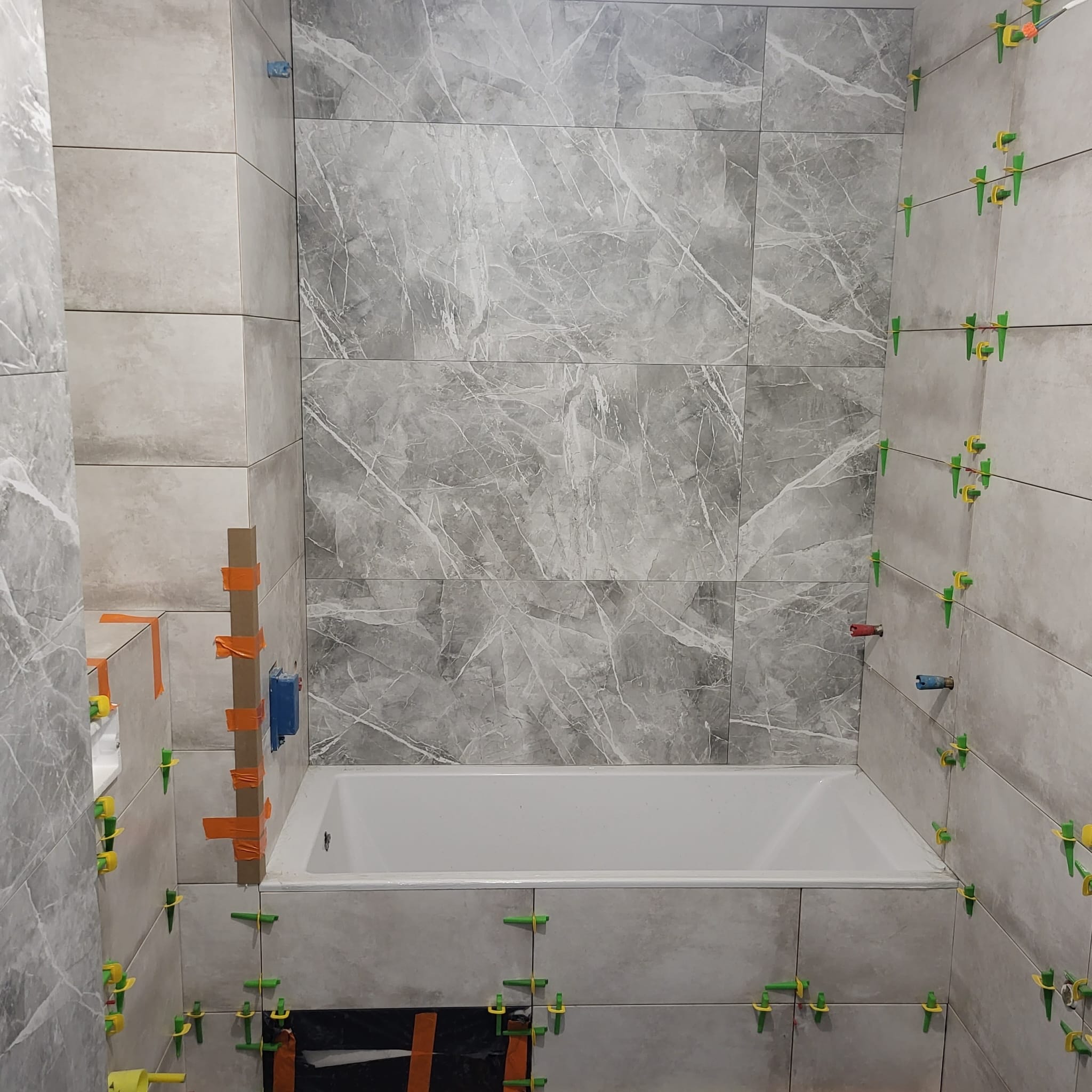 Biała wanna obudowana szarymi płytkami imitującymi marmur, z widocznymi krzyżykami dystansowymi i poziomowaniem, częściowo wykończona ściana z szarych płytek.