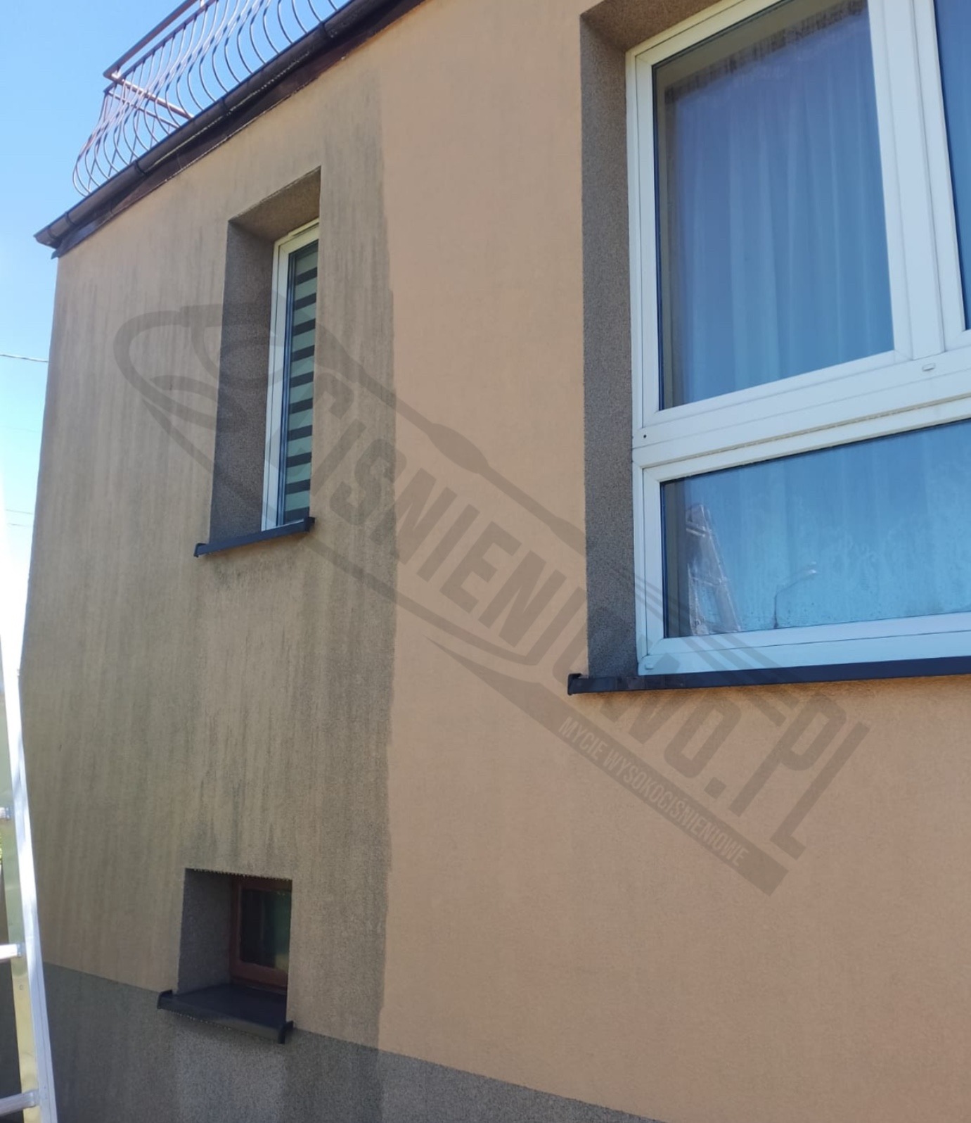 Fragment elewacji budynku z widocznym efektem mycia: czysta, jasna część kontrastuje z brudną, ciemniejszą powierzchnią. Widoczne okna z białymi ramami i balkon na górze. Na elewacji widoczne ślady...