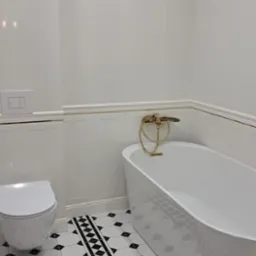 Biała łazienka z wolnostojącą wanną i złotą armaturą, ściany wyłożone białymi płytkami z delikatnym pionowym wzorem, podłoga w biało-czarne geometryczne wzory.