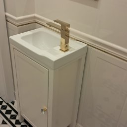 Best- Fliz Katowice - Umywalka z szafką w stylu retro, złota bateria, czarno-biała podłoga w karo, dekoracyjne listwy ścienne z cienką złotą linią.