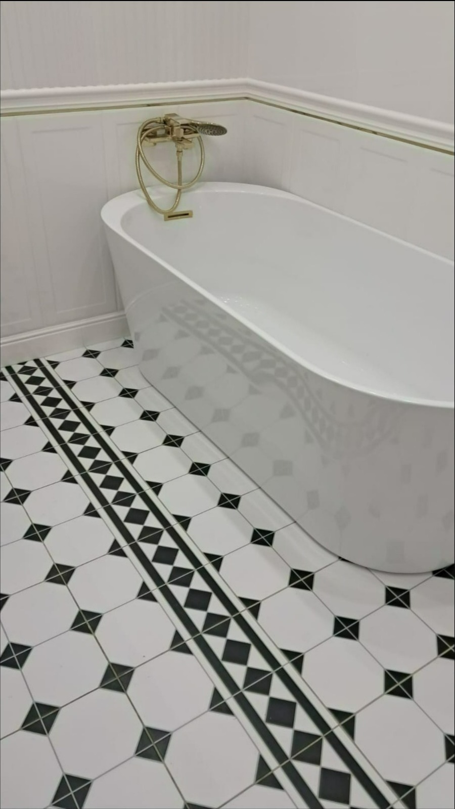 Elegancka biała wanna wolnostojąca z złotą armaturą na tle białych paneli ściennych z delikatnym złotym akcentem i podłogi z czarno-białych płytek w geometryczny wzór.