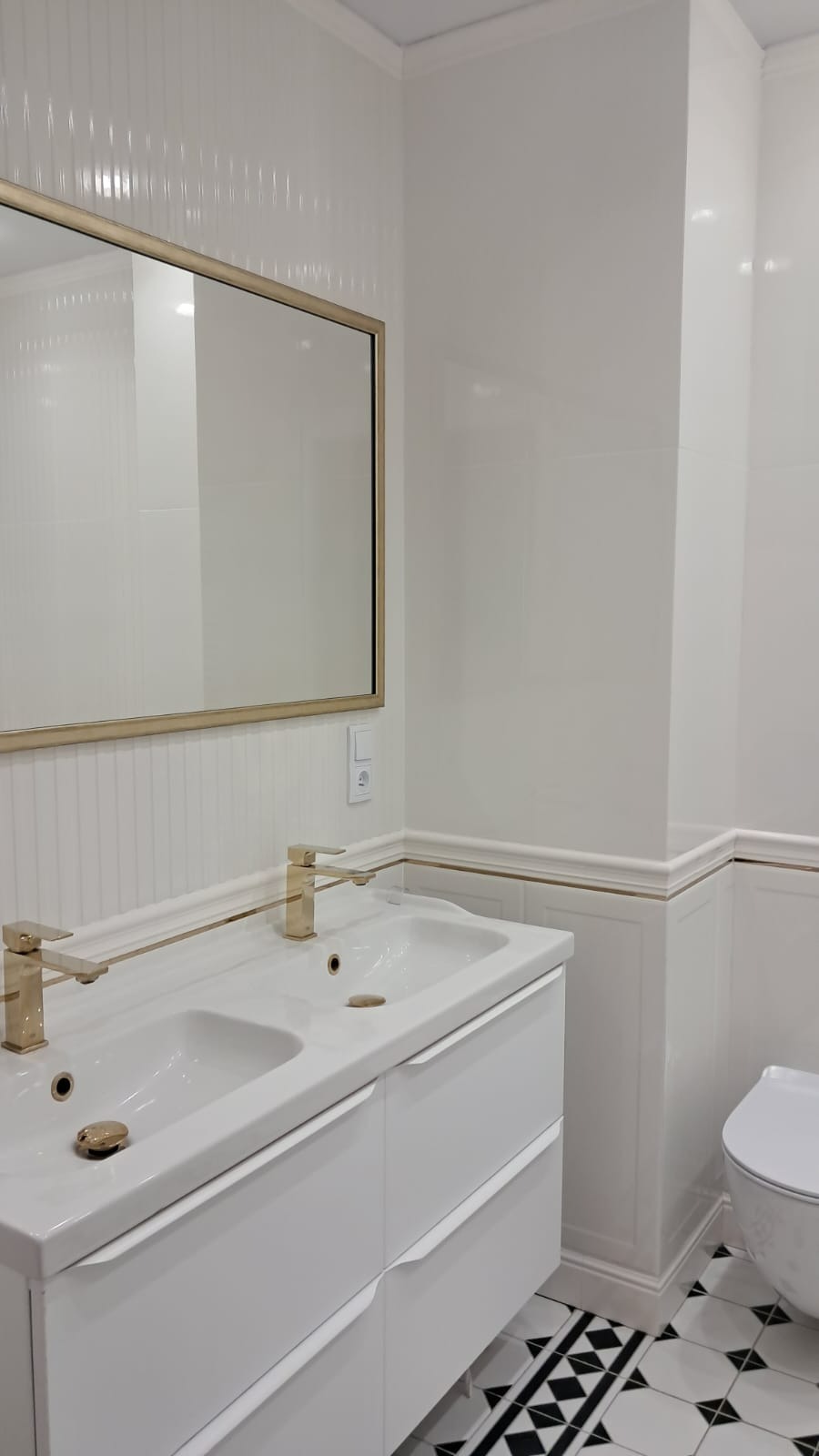Biała łazienka z podwójną umywalką, złotymi bateriami i lustrem w złotej ramie, wykończona białymi płytkami z delikatnym wzorem, czarno-biała podłoga z geometrycznym wzorem.