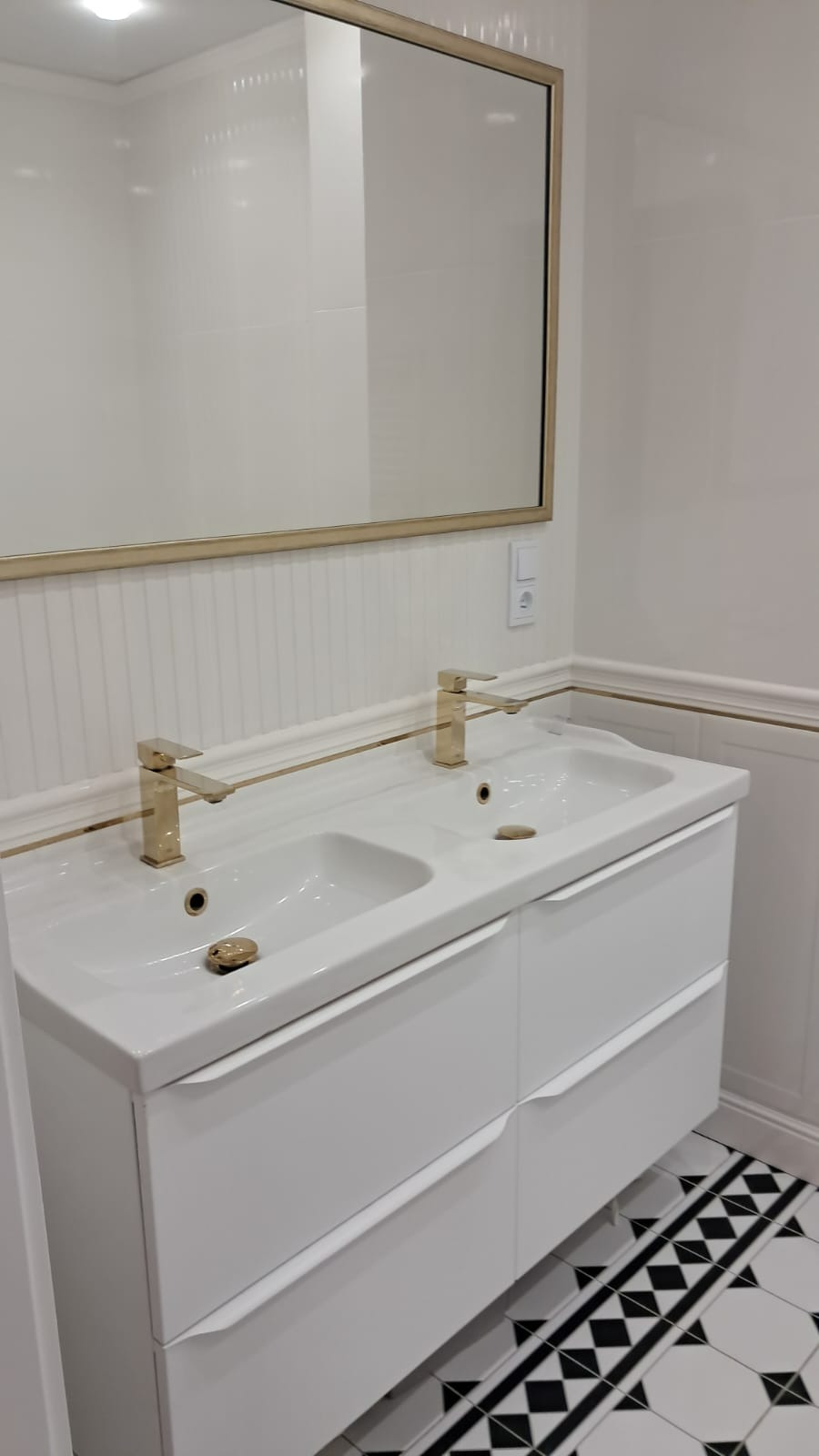 Biała umywalka podwójna z szafką i złotymi bateriami w nowoczesnej łazience z białą glazurą i czarno-białą podłogą w geometryczny wzór.