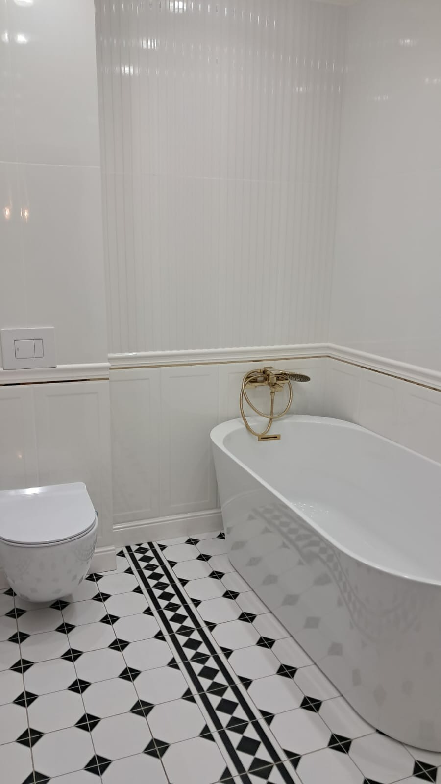 Biała łazienka z wolnostojącą wanną i złotą armaturą, ściany wyłożone białymi płytkami z delikatnym pionowym wzorem, podłoga w biało-czarne geometryczne wzory.