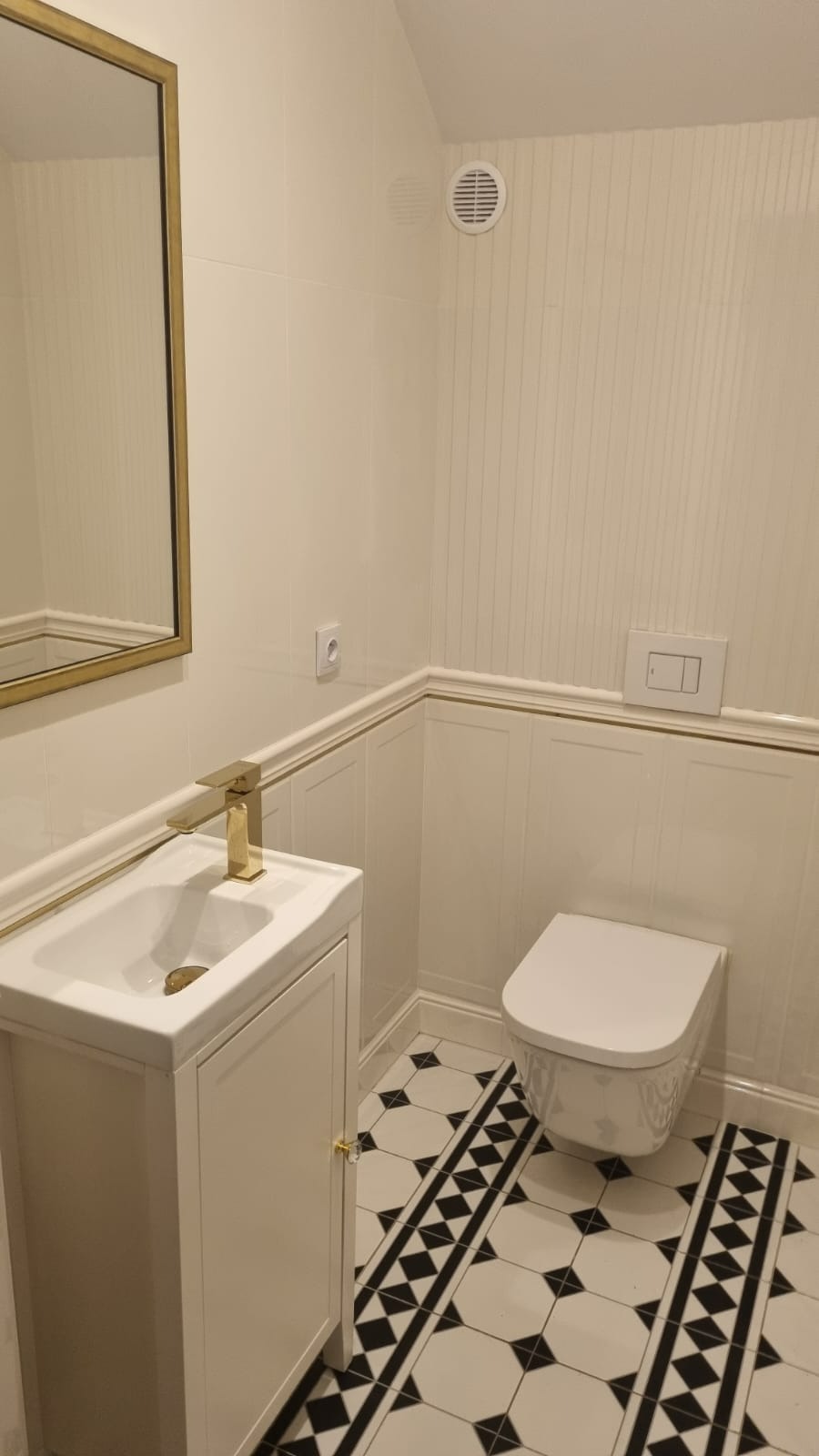 Biała łazienka z czarno-białą podłogą w romby, złota armatura i rama lustra, dekoracyjne panele ścienne i wisząca toaleta.