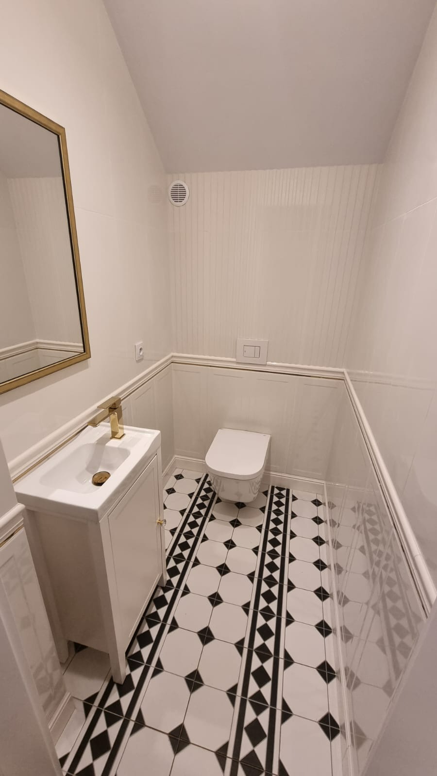 Nowoczesna toaleta z białą umywalką na szafce z złotą armaturą, lustrem w złotej ramie i czarno-białą podłogą w geometryczny wzór.