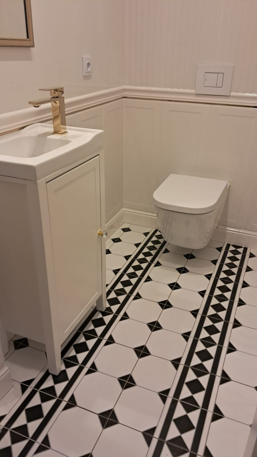 Nowoczesna łazienka z białymi panelami ściennymi, podwieszaną toaletą, szafką umywalkową z złotą armaturą i charakterystyczną czarno-białą podłogą w geometryczne wzory.