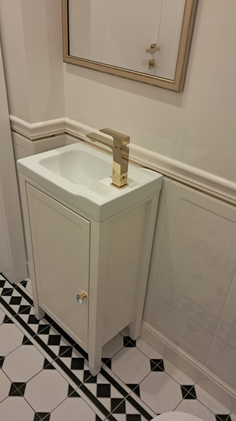 Umywalka z szafką w stylu retro, złota bateria, czarno-biała podłoga w karo, dekoracyjne listwy ścienne z cienką złotą linią.