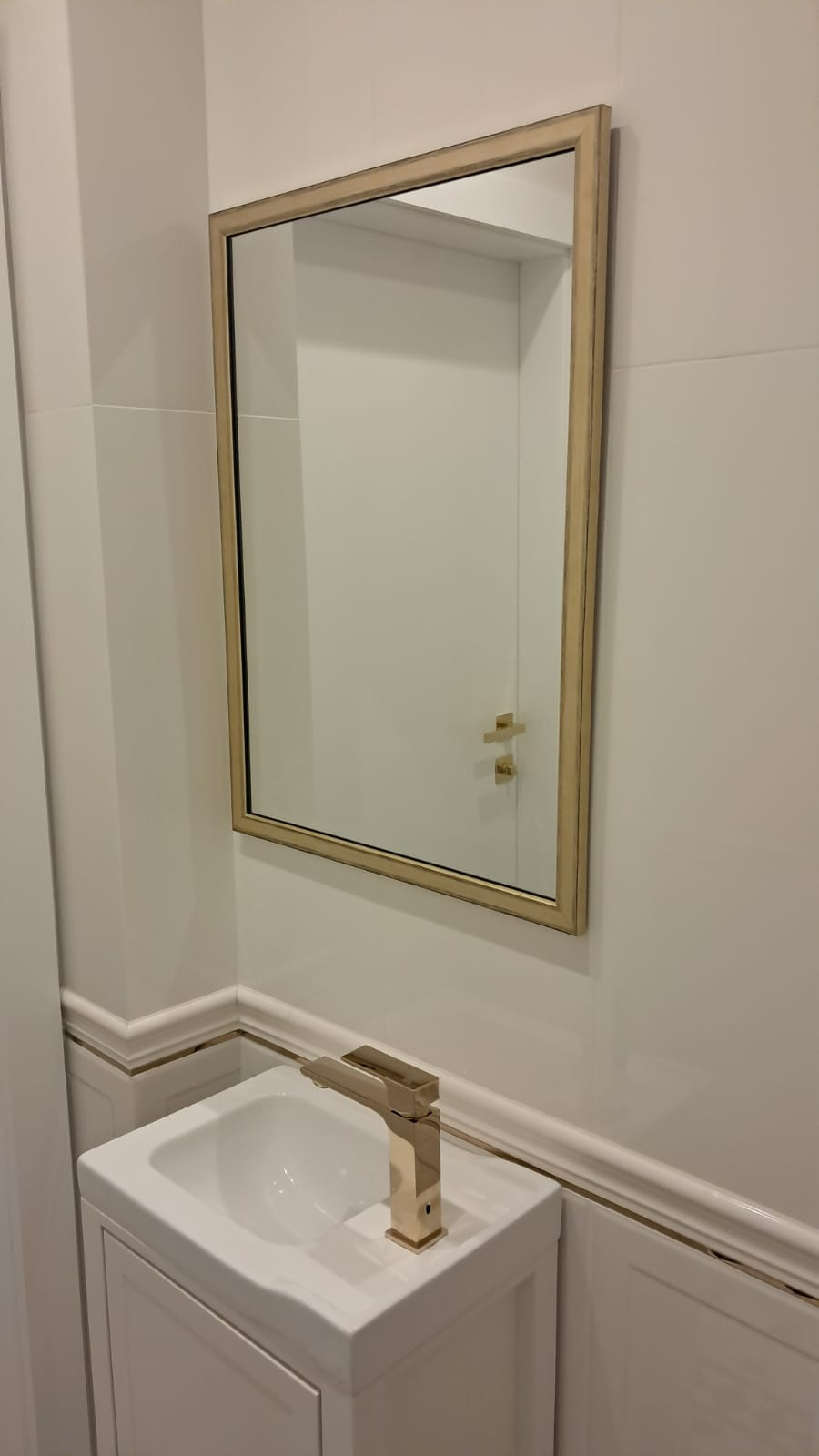 Biała umywalka z szafką i złotą baterią, nad nią lustro w złotej ramie, widoczne białe płytki z dekoracyjnym paskiem.