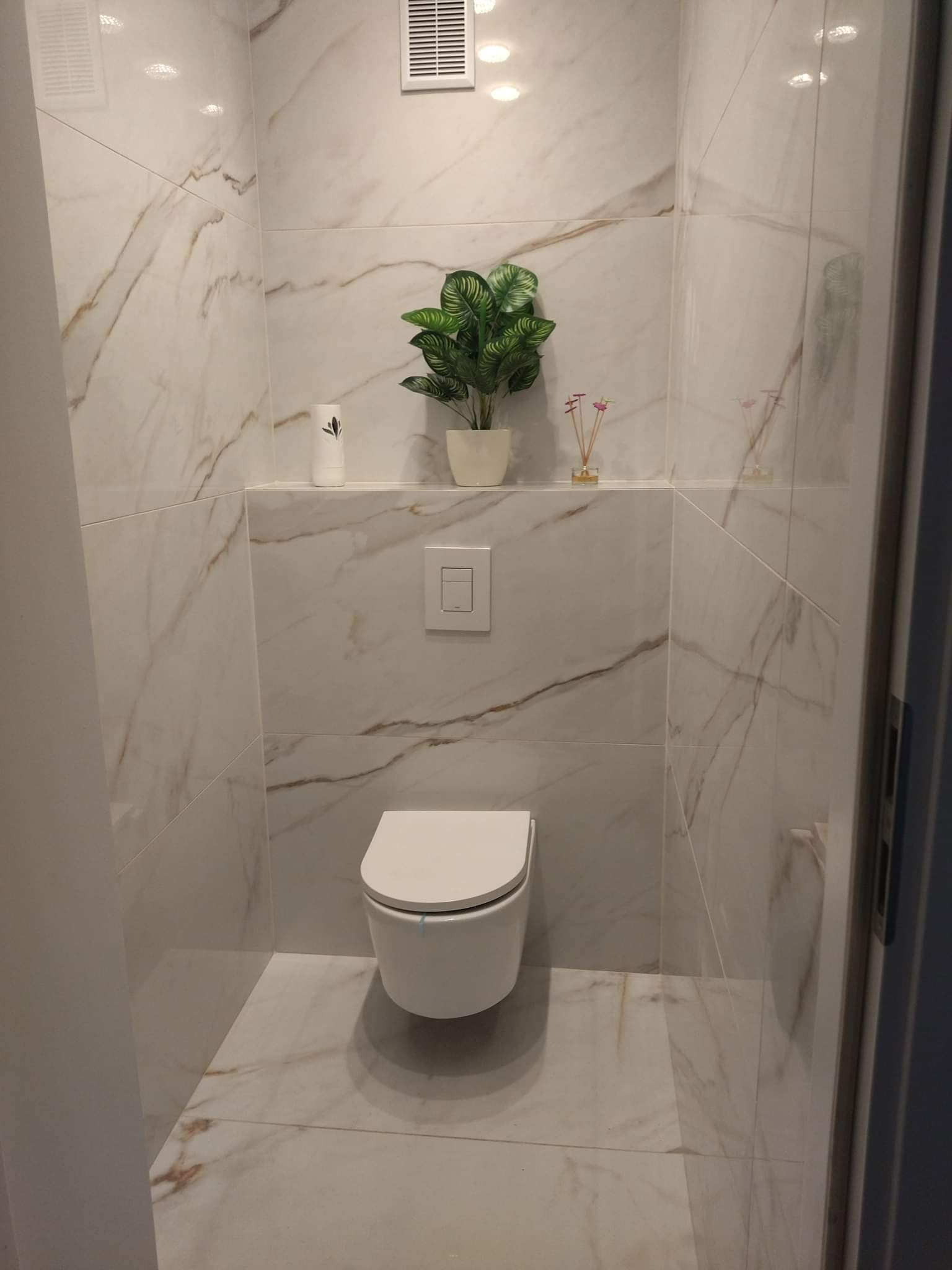 Nowoczesna toaleta w wykończonej łazience, marmurowe płytki z brązowymi żyłami na ścianach i podłodze, wisząca muszla klozetowa, półka z dekoracjami: sztuczna roślina w doniczce i dyfuzor zapachowy.