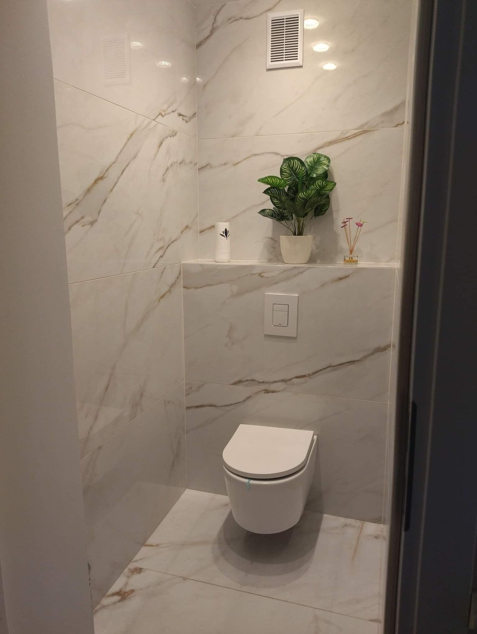 Nowoczesna toaleta podwieszana w wykończonej łazience, białe płytki imitujące marmur z subtelnym złotym żyłkowaniem, dekoracyjna roślina w doniczce na półce, minimalistyczny design.