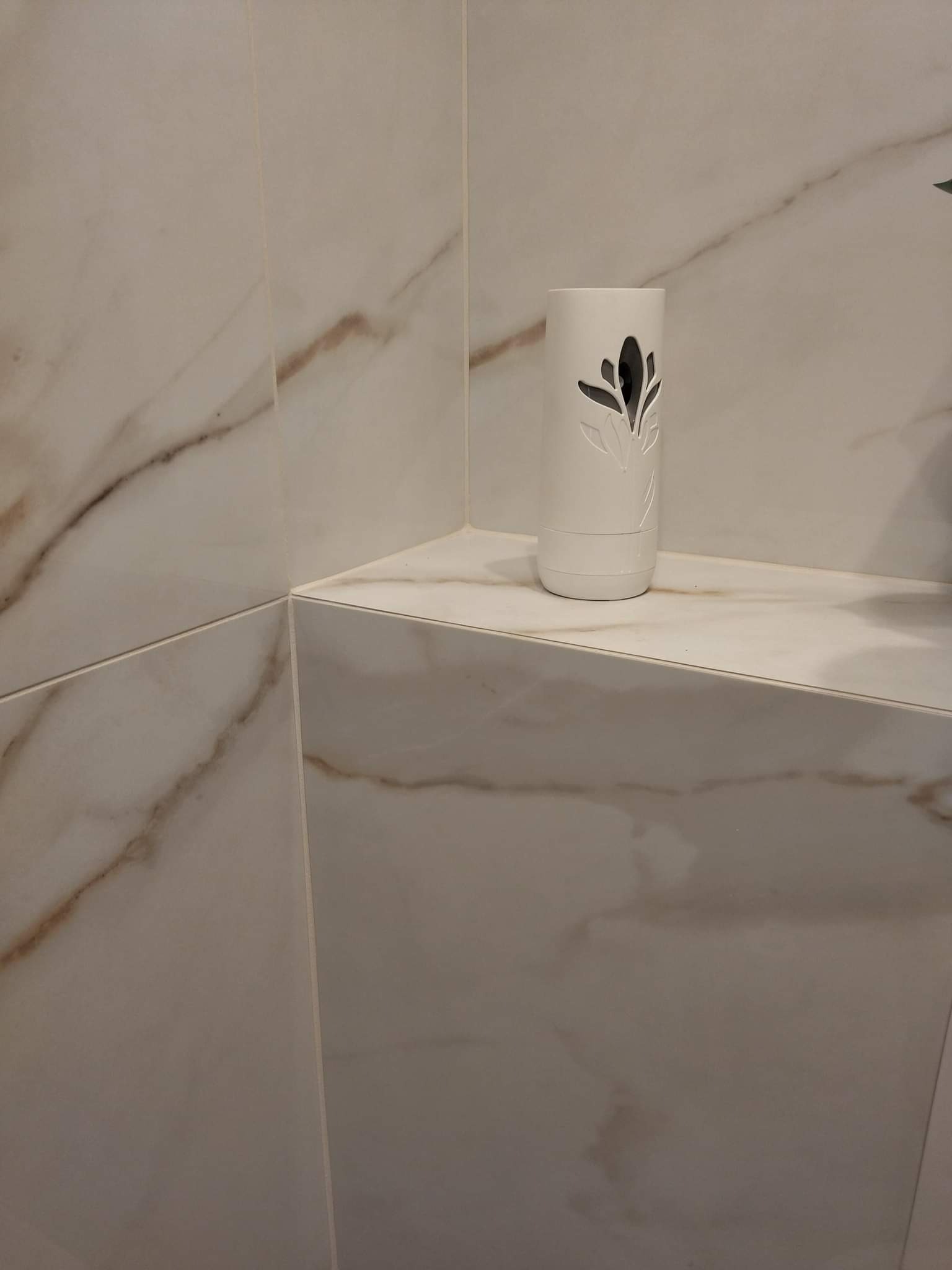 Białe płytki imitujące marmur z brązowymi żyłami, ułożone na ścianie i półce w rogu łazienki. Na półce stoi biały odświeżacz powietrza z wyciętym motywem roślinnym.