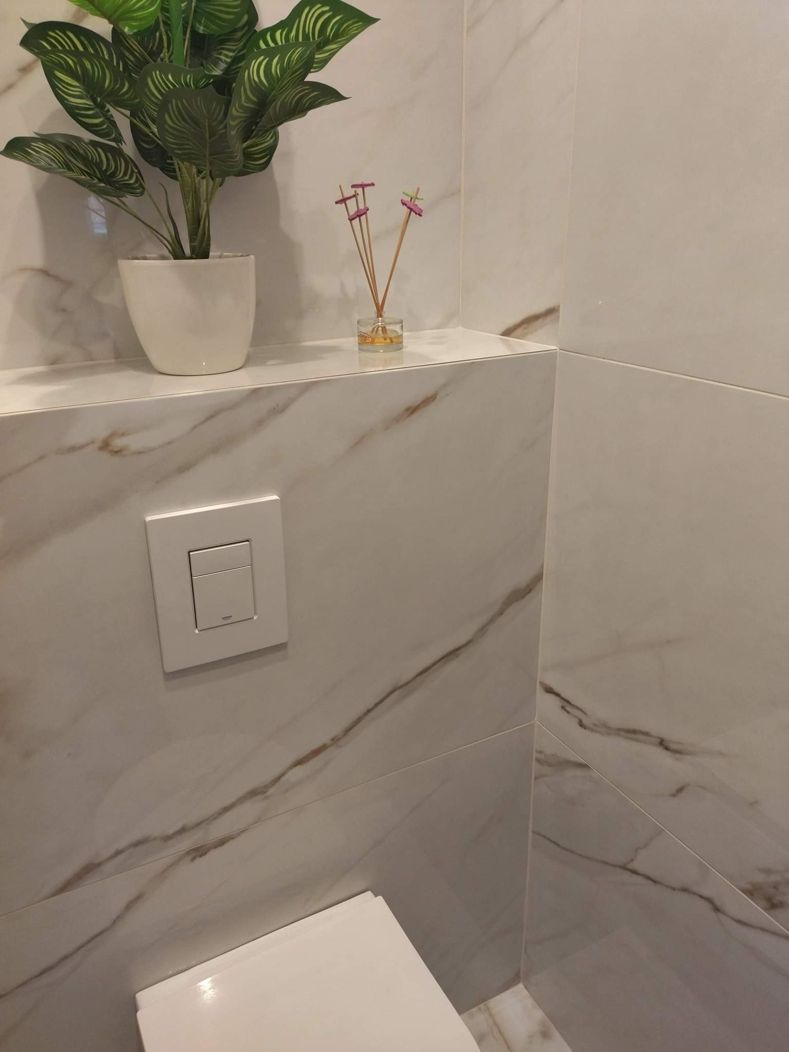 Nowoczesna toaleta z marmurowymi płytkami, ukryta spłuczka, dekoracyjna roślina w białej doniczce i zapachowe patyczki na półce.