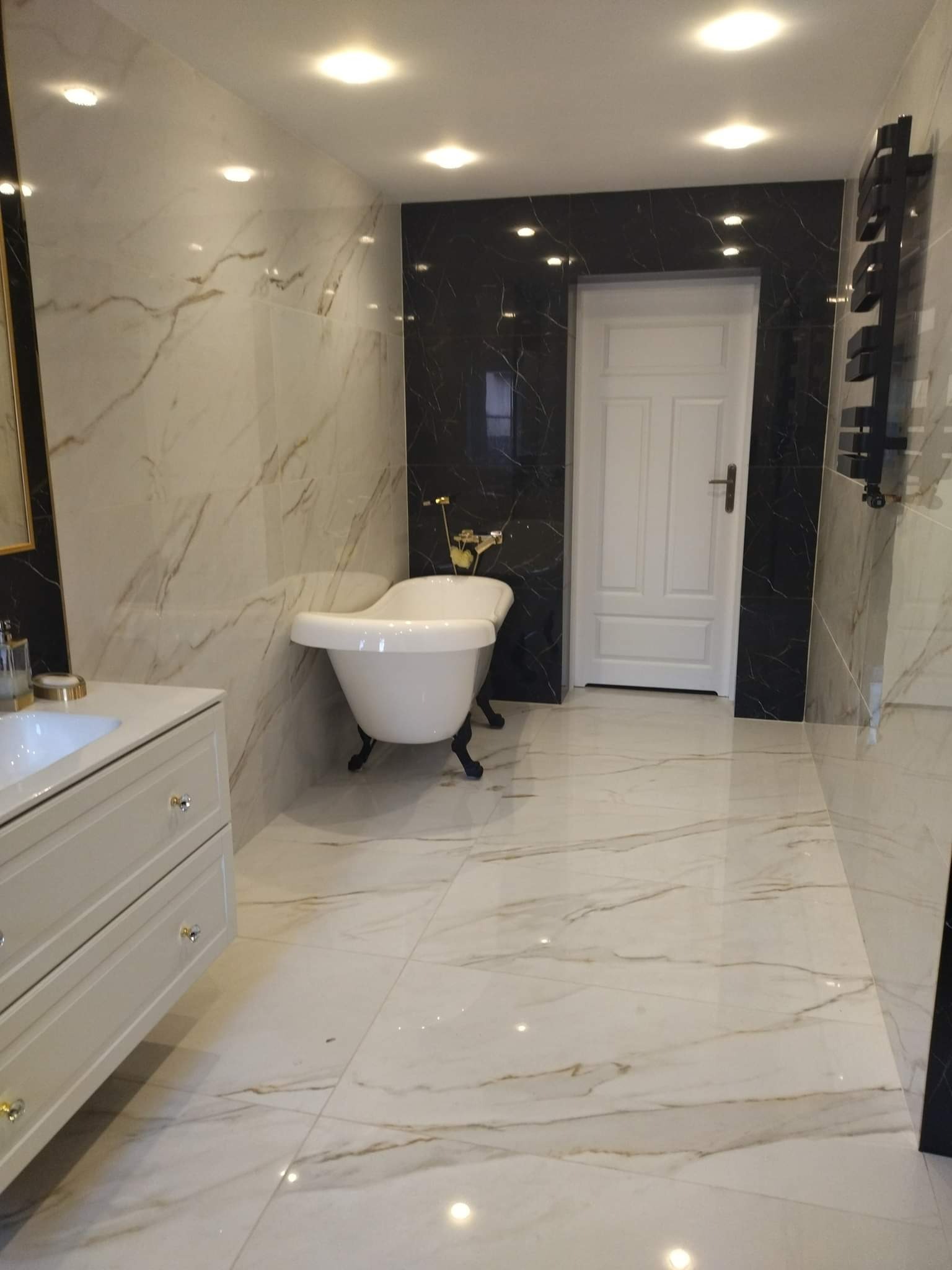 Elegancka łazienka z wolnostojącą wanną na lwich nóżkach, marmurowe płytki w odcieniach bieli i złota, czarne akcenty płytek i grzejnika drabinkowego, oświetlenie punktowe.