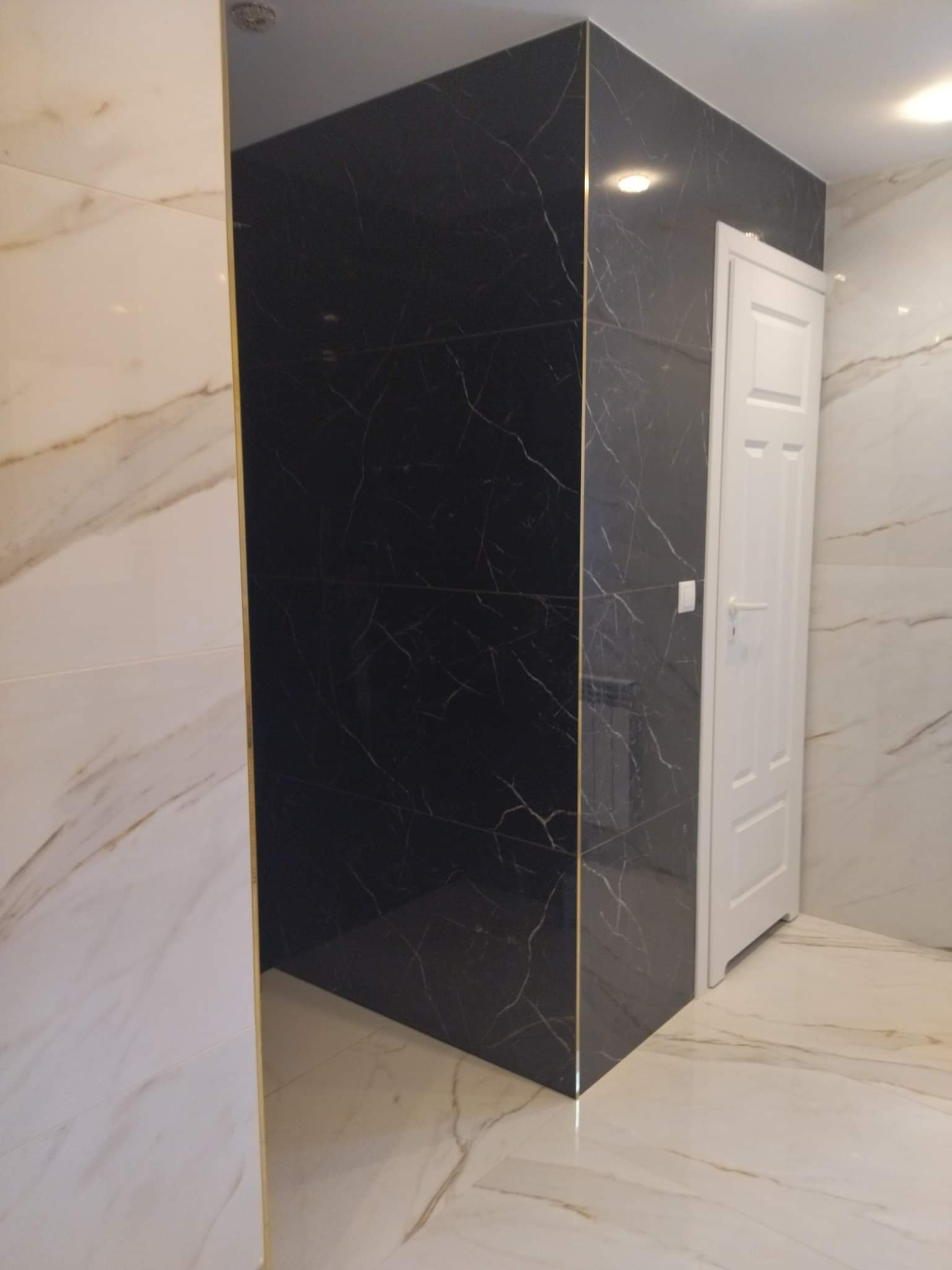 Wykończona łazienka z czarnymi płytkami imitującymi marmur z delikatnym złotym akcentem na krawędziach, kontrastującymi z jasnymi płytkami marmurowymi na podłodze i ścianach oraz białą, panelową...