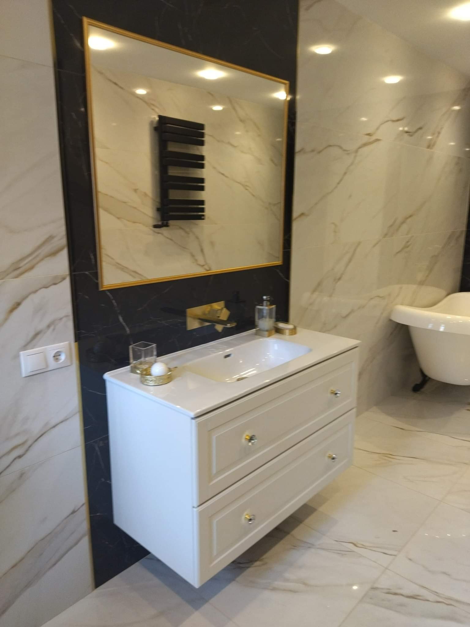 Elegancka łazienka z marmurowymi płytkami, białą szafką pod umywalką z dwoma szufladami i złotymi gałkami, oraz owalną wanną na nóżkach w tle. Na ścianie wisi lustro w złotej ramie, w którym odbija...