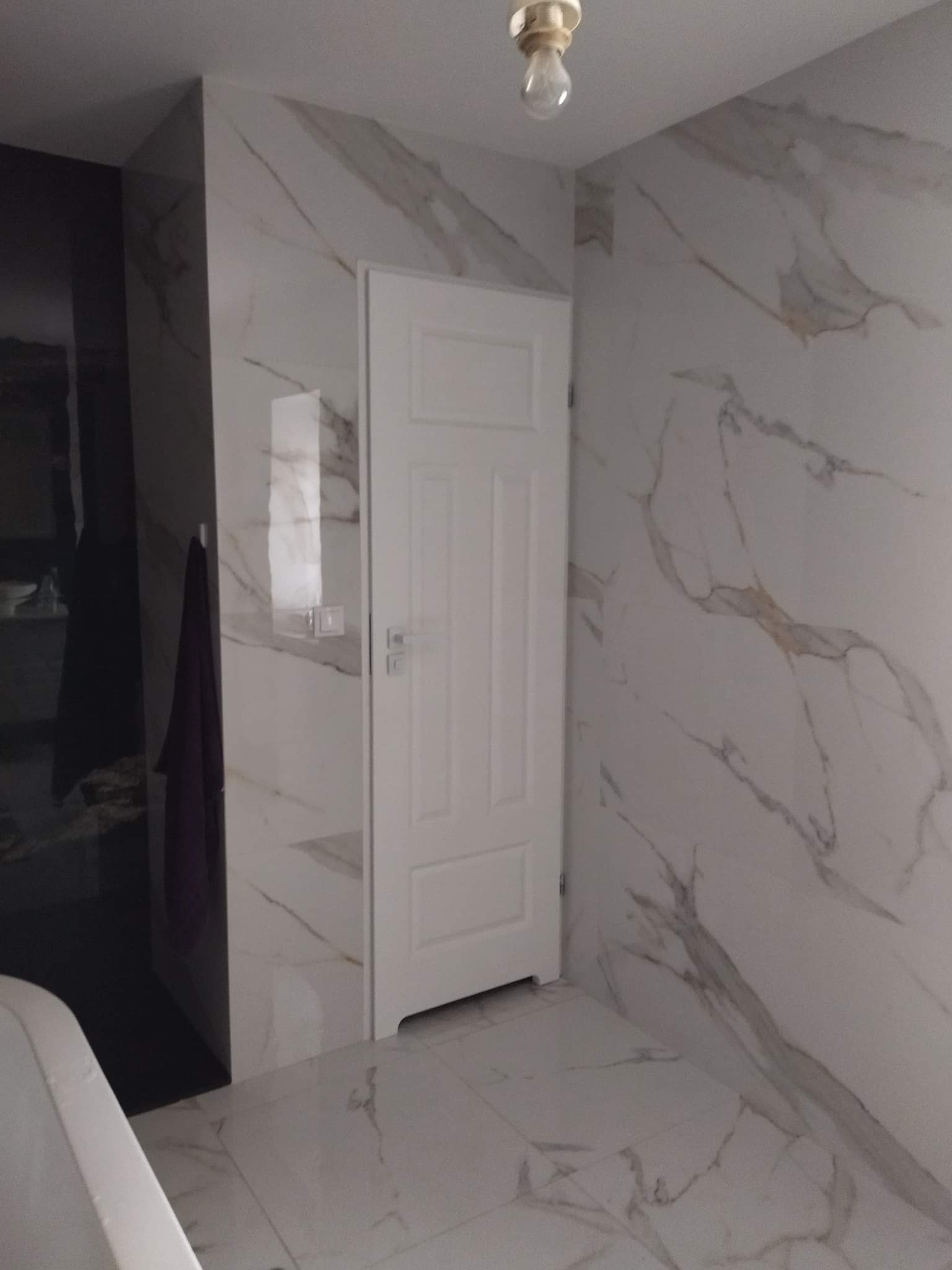 Wykończona łazienka z białymi płytkami imitującymi marmur na ścianach i podłodze, białe drzwi wewnętrzne, widoczna część wanny i wiszący ręcznik.